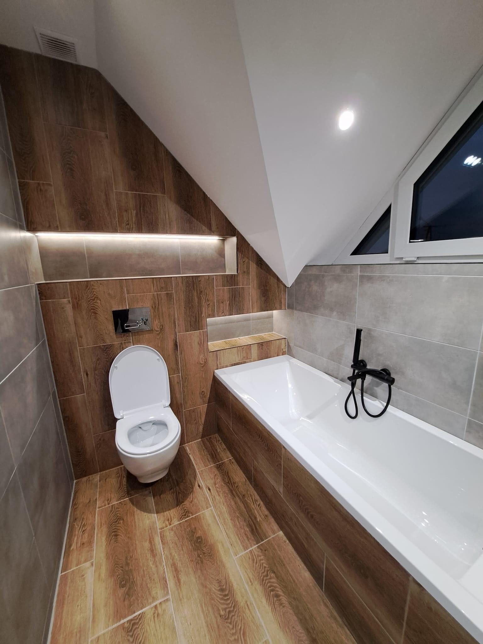 Ukończona łazienka na poddaszu ze skośnym sufitem, wykończona płytkami imitującymi drewno i szare płytki, z białą wanną i czarną armaturą oraz podświetlaną wnęką nad toaletą.