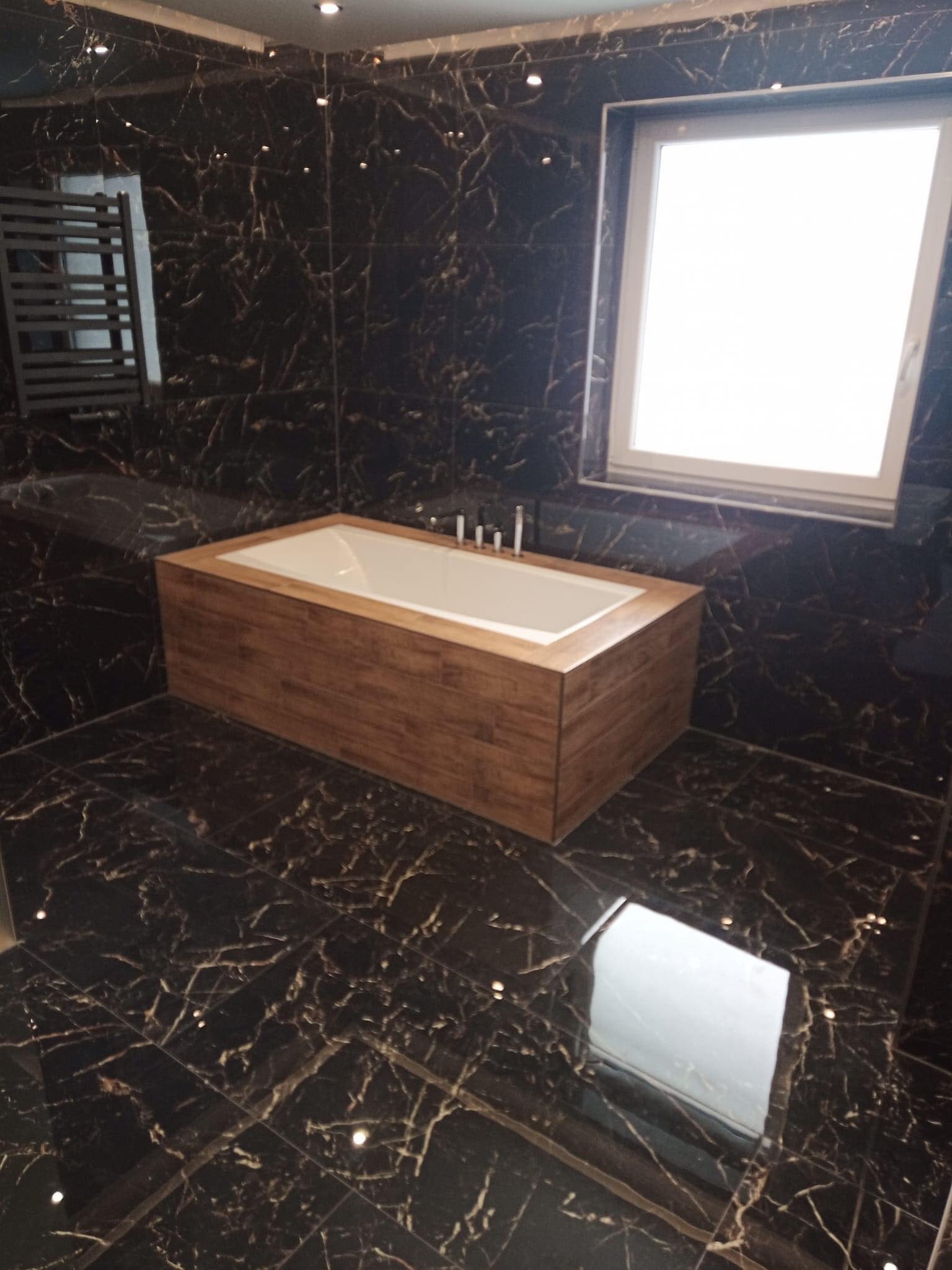 Nowoczesna łazienka z wanną obudowaną drewnem, czarne marmurowe płytki na ścianach i podłodze, okno z białymi ramami w tle.