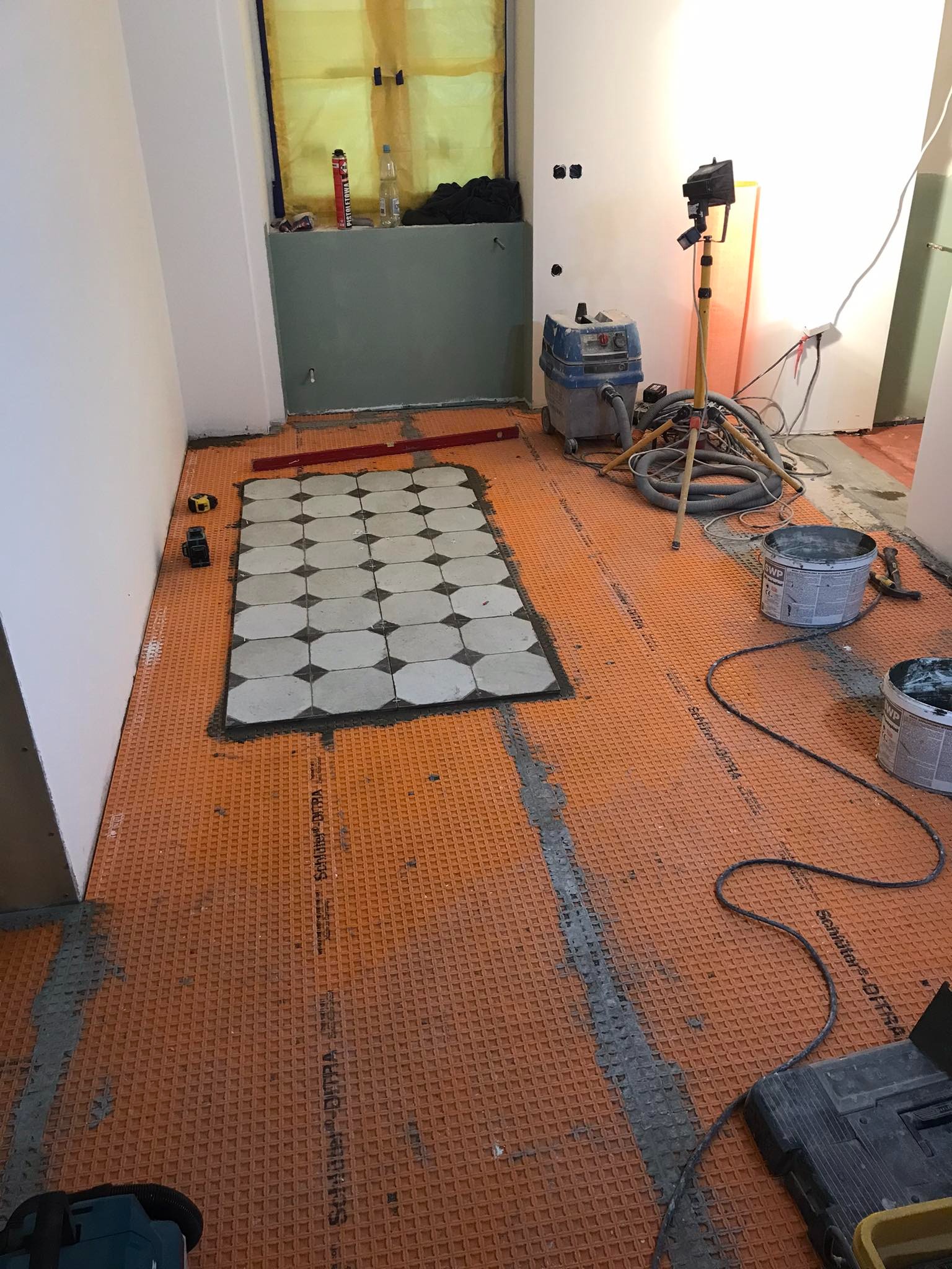 Przygotowanie podłoża z pomarańczowej membrany drenażowej Schluter Ditra pod układanie płytek podłogowych, widoczny fragment ułożonych płytek w geometryczny wzór, narzędzia i wiadro z zaprawą w tle.