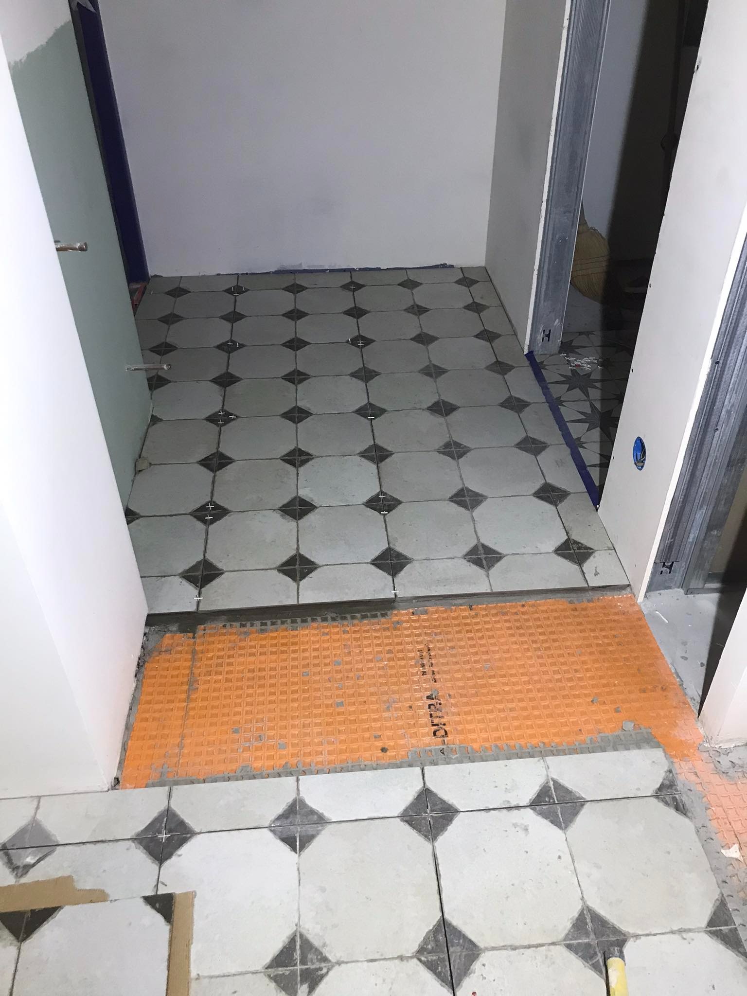 Podłoga w trakcie remontu z częściowo ułożonymi, szaro-białymi płytkami w romby i pomarańczową membraną uszczelniającą DITRA, widoczne ślady zaprawy.
