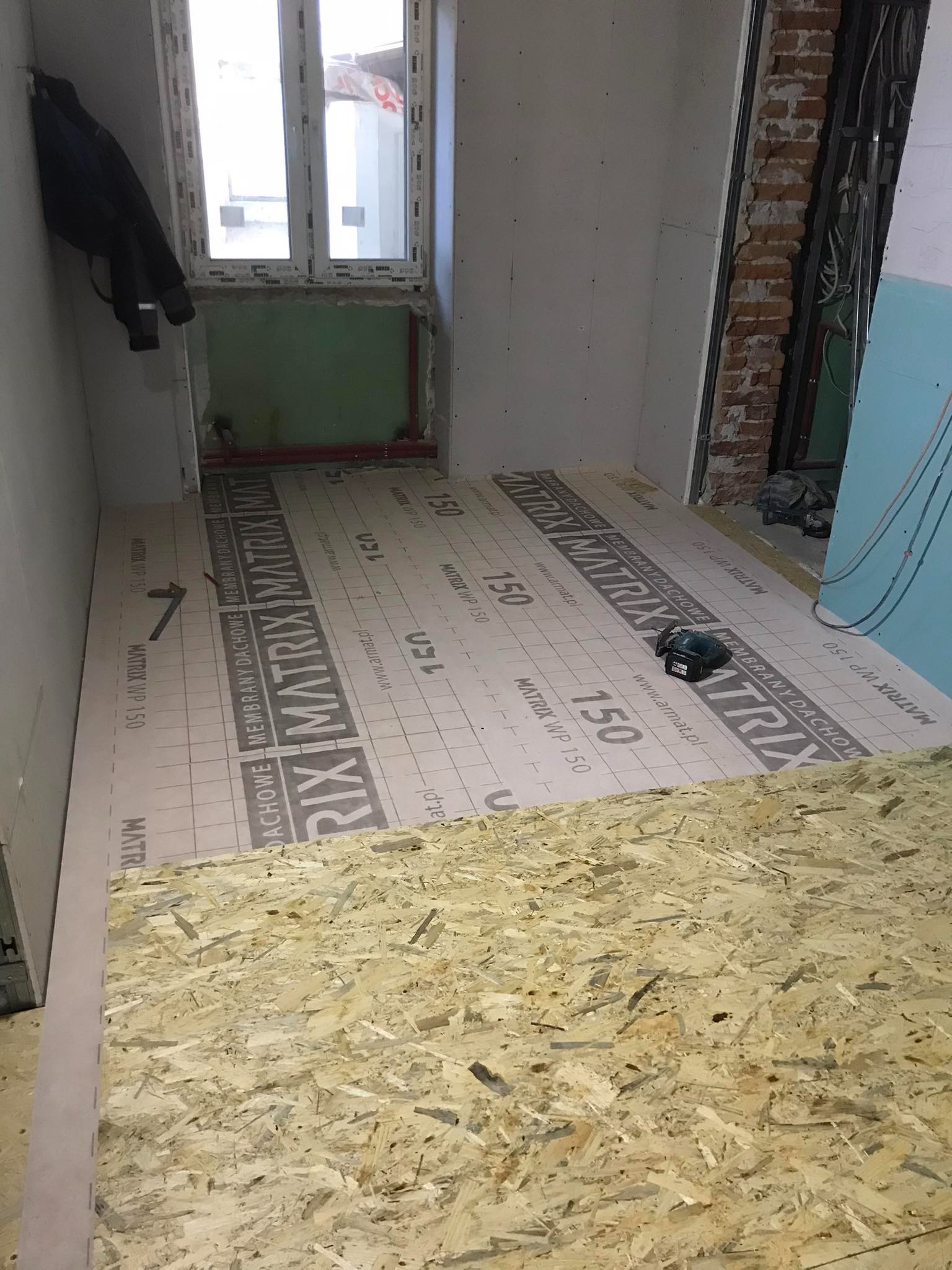 Przygotowanie podłoża w pomieszczeniu: płyty OSB na membranie dachowej Matrix WP 150, widoczne fragmenty instalacji i ścian w trakcie prac wykończeniowych, narzędzie akumulatorowe na membranie.