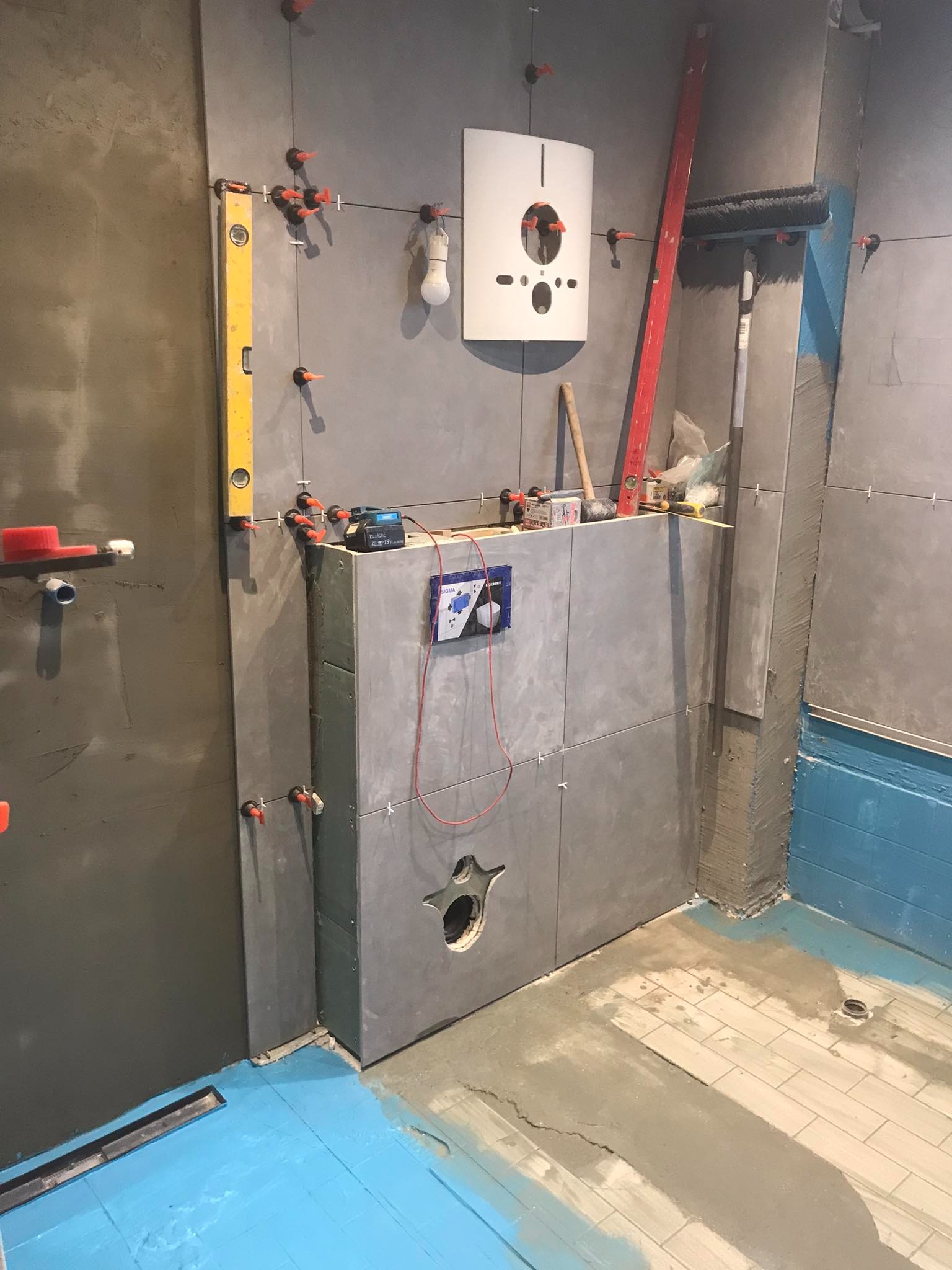 Szare płytki ceramiczne układane na ścianie w łazience, widoczne narzędzia glazurnicze, poziomica, system poziomowania płytek, instalacja podtynkowa WC w trakcie montażu, zabezpieczenie podłogi...
