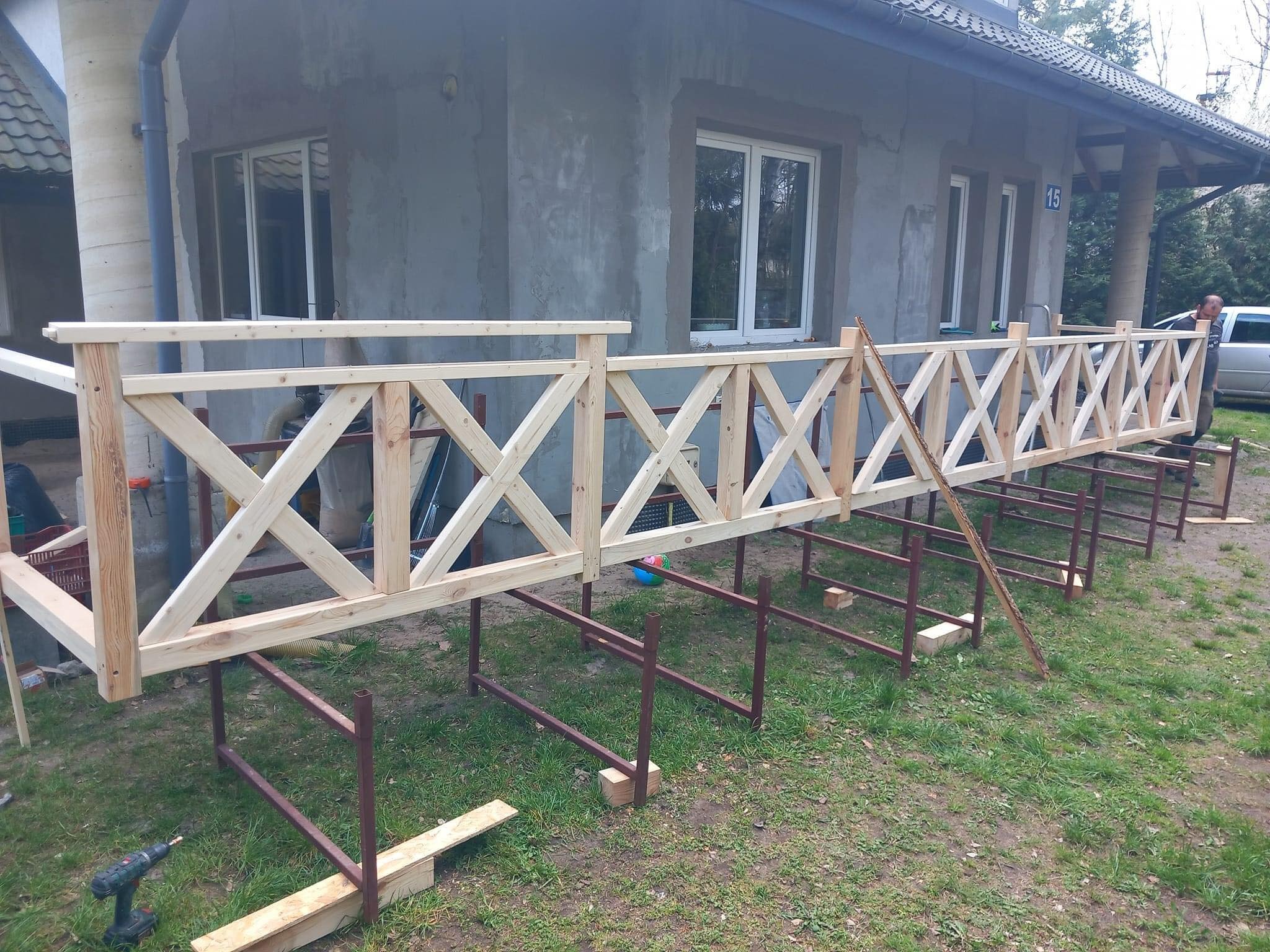 Surowa, drewniana balustrada z krzyżowymi wzorami, ustawiona na metalowych stelażach przed szarym domem w trakcie montażu, widoczna wiertarka na trawie.