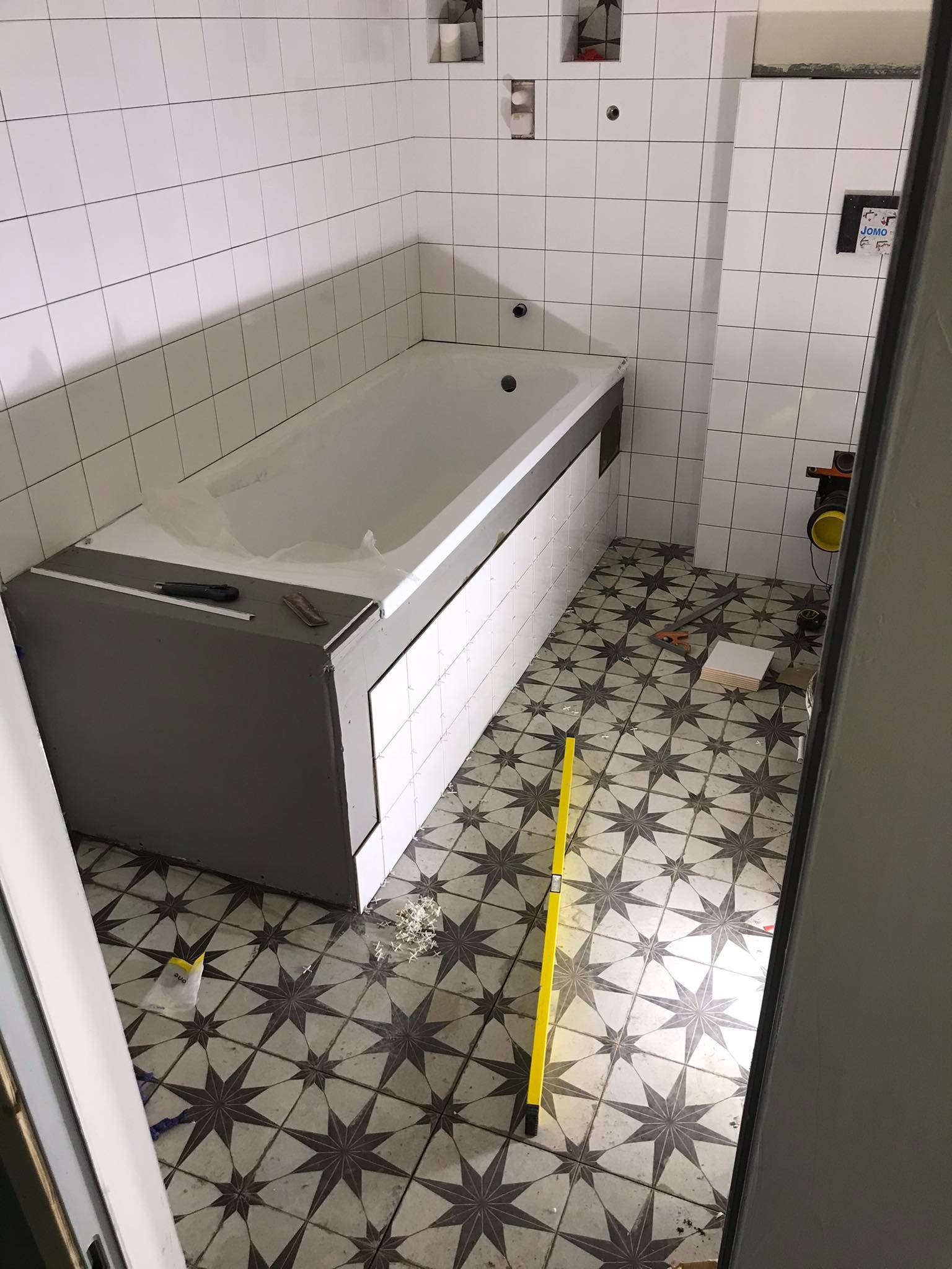 Biała wanna z obudową w trakcie wykańczania, widoczne narzędzia glazurnicze, poziomica na podłodze z wzorem w gwiazdy, białe płytki na ścianach.