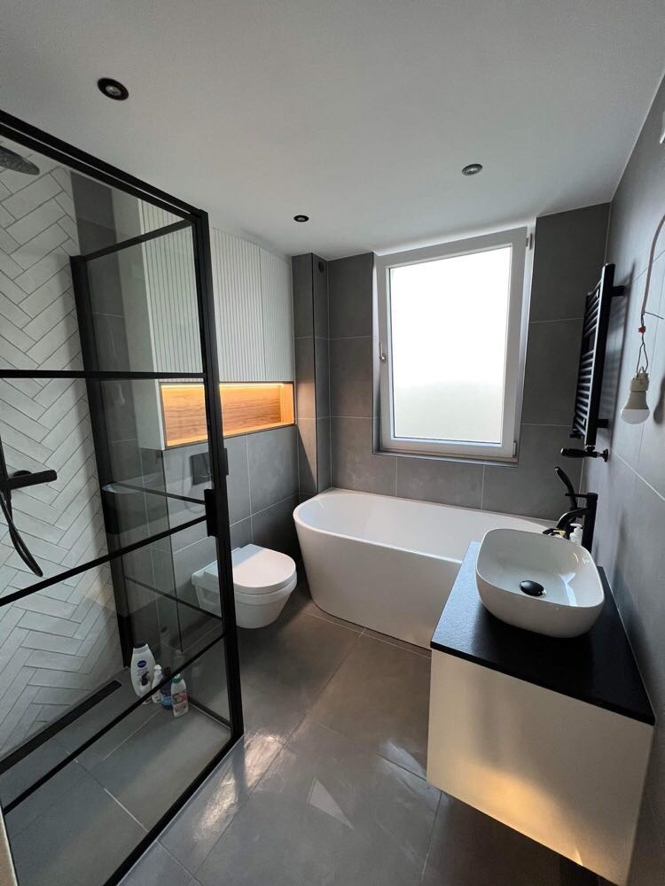 Nowoczesna łazienka z szarą glazurą, kabiną prysznicową z czarnymi profilami, wolnostojącą wanną, umywalką nablatową na białej szafce z czarnym blatem i podświetlaną wnęką nad toaletą.