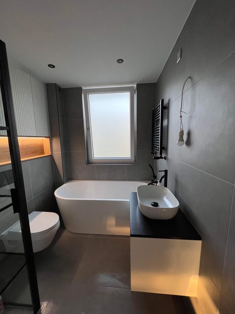 Nowoczesna łazienka z wolnostojącą wanną, podwieszaną toaletą, umywalką nablatową na czarnym blacie i grzejnikiem drabinkowym, oświetlona wiszącą żarówką na kablu.