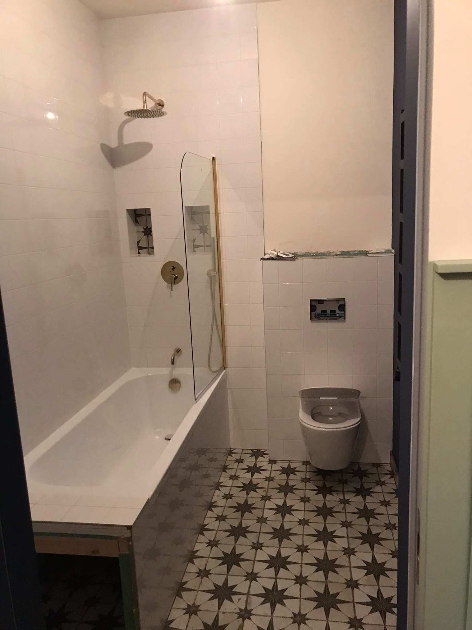 Remontowana łazienka z białą wanną, złotą armaturą, szklanym parawanem, wzorzystą podłogą i wiszącą toaletą przed montażem panelu.