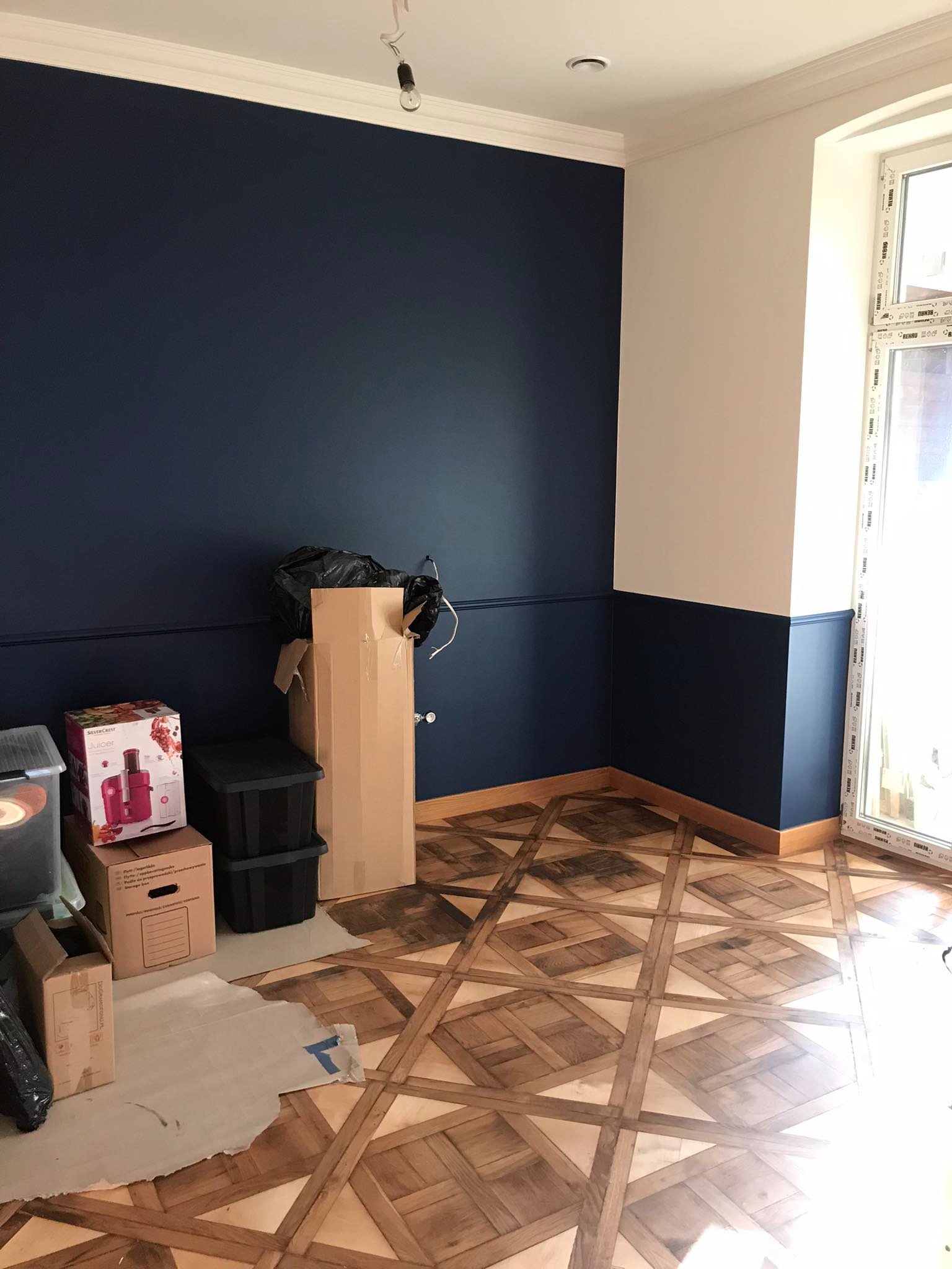 Pokój po malowaniu z ciemnoniebieską ścianą do połowy wysokości, białą górną częścią i ozdobną listwą, parkiet w kwadraty, kartony i pudła na podłodze, okno z białymi ramami.