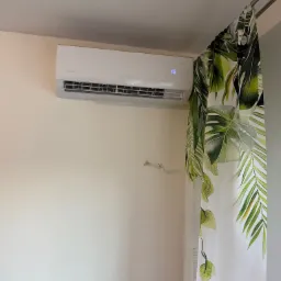 Biały klimatyzator Rotenso zamontowany na ścianie w pomieszczeniu, obok zasłony z motywem liści tropikalnych, wyświetlający temperaturę 16 stopni.