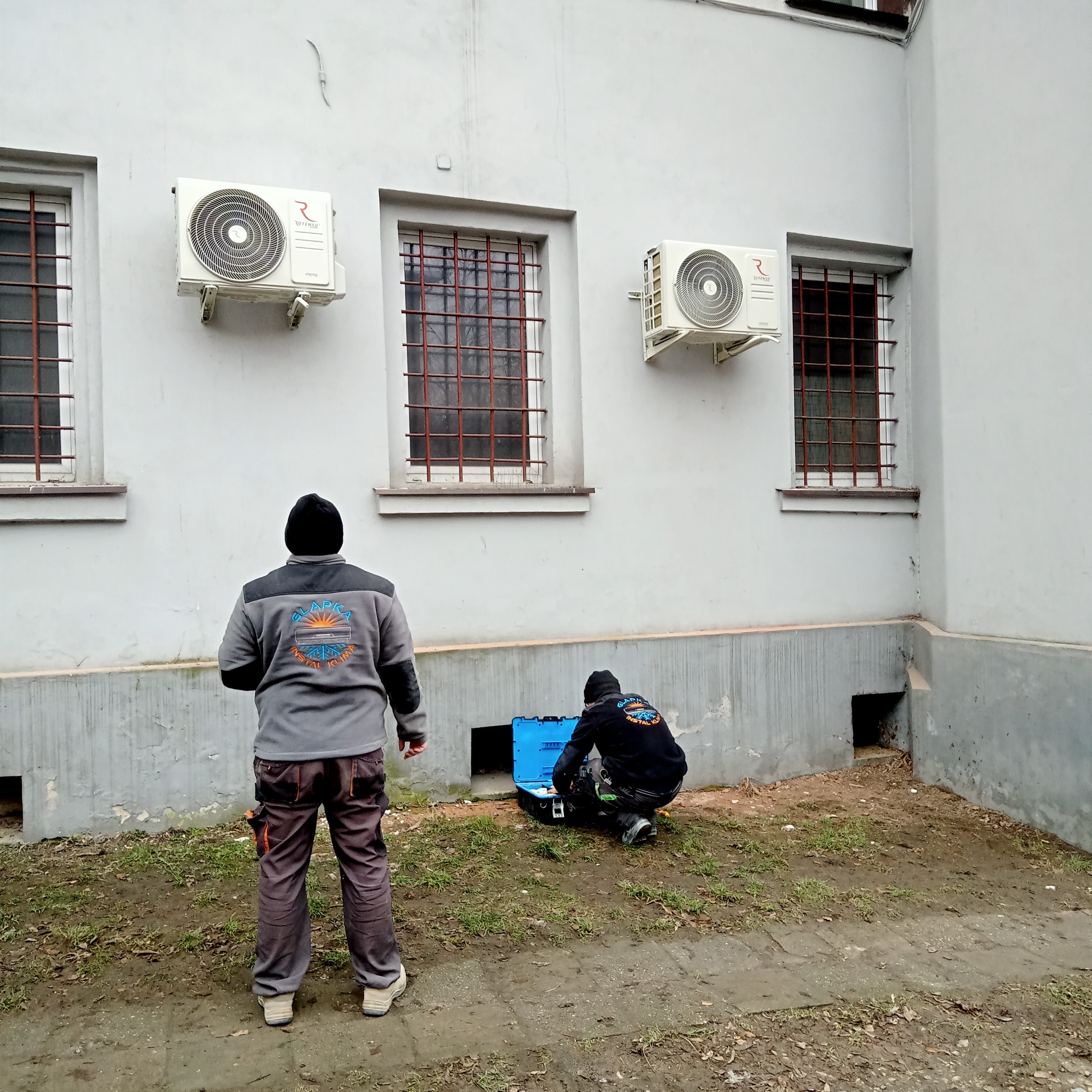 Dwóch instalatorów przy budynku z zamontowanymi klimatyzatorami, jeden stoi, drugi pracuje przy niebieskiej skrzynce z narzędziami.