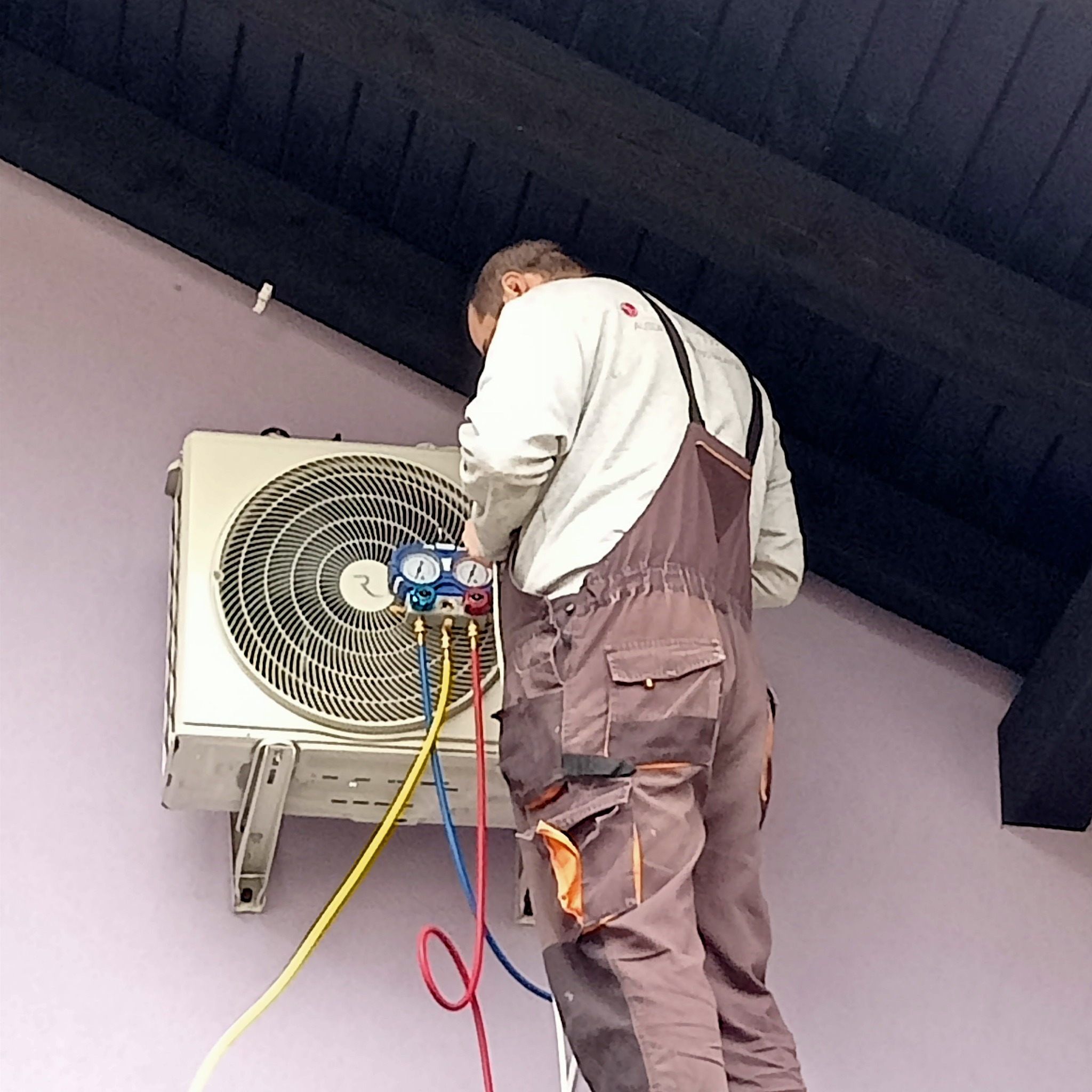 Instalator w szarym kombinezonie podłączony do zewnętrznej jednostki klimatyzacji na jasnofioletowej ścianie, używa manometru z kolorowymi wężami.