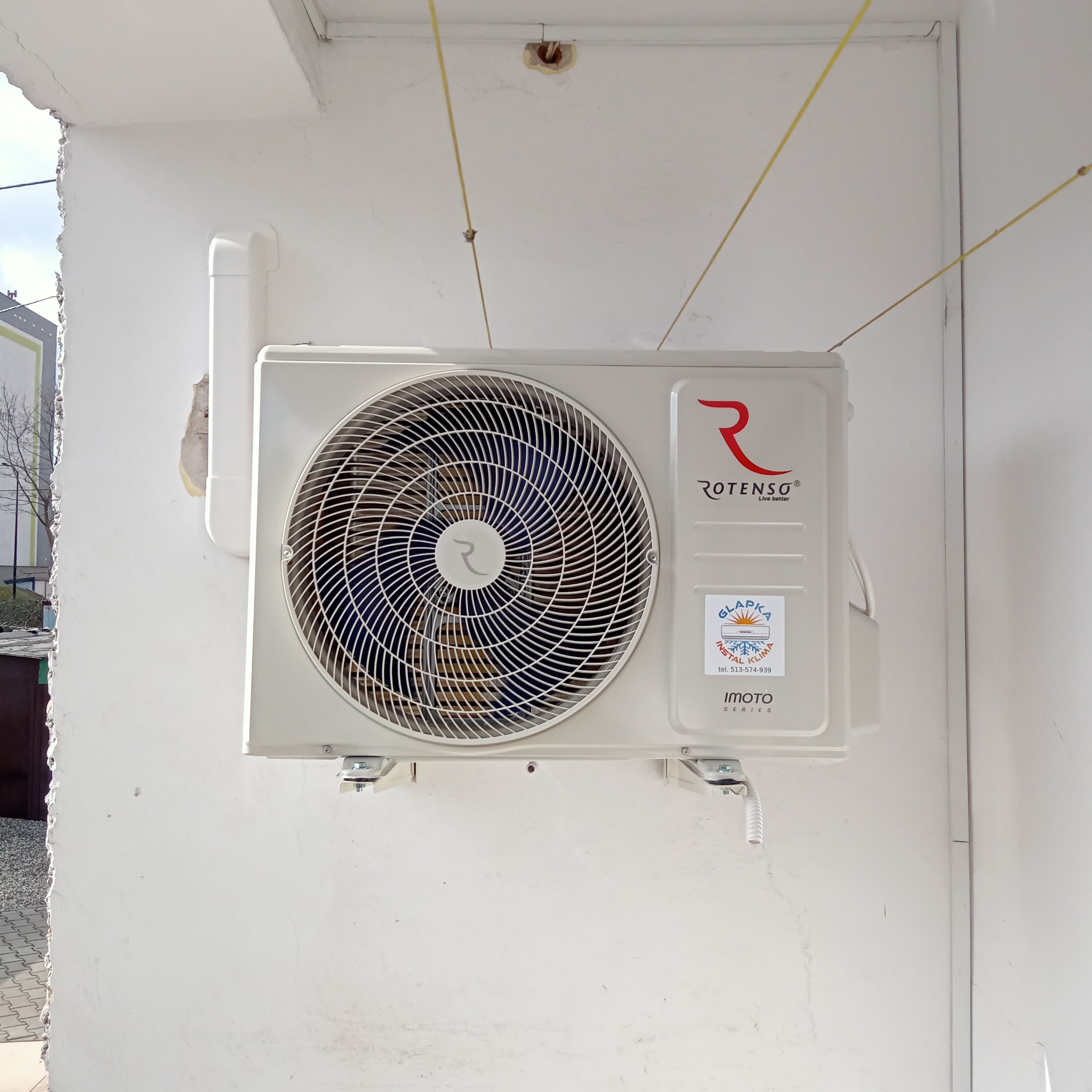 Biała jednostka zewnętrzna klimatyzacji Rotenso IMOTO Series zamontowana na białej ścianie budynku, widoczne przewody i naklejka firmy instalacyjnej.