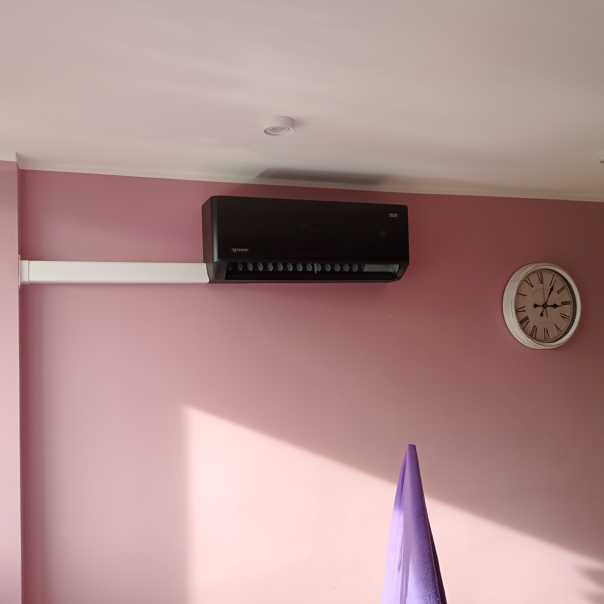 Czarny klimatyzator Rotenso Teta zamontowany na różowej ścianie, z białą listwą maskującą przewody i zegarem w stylu vintage.