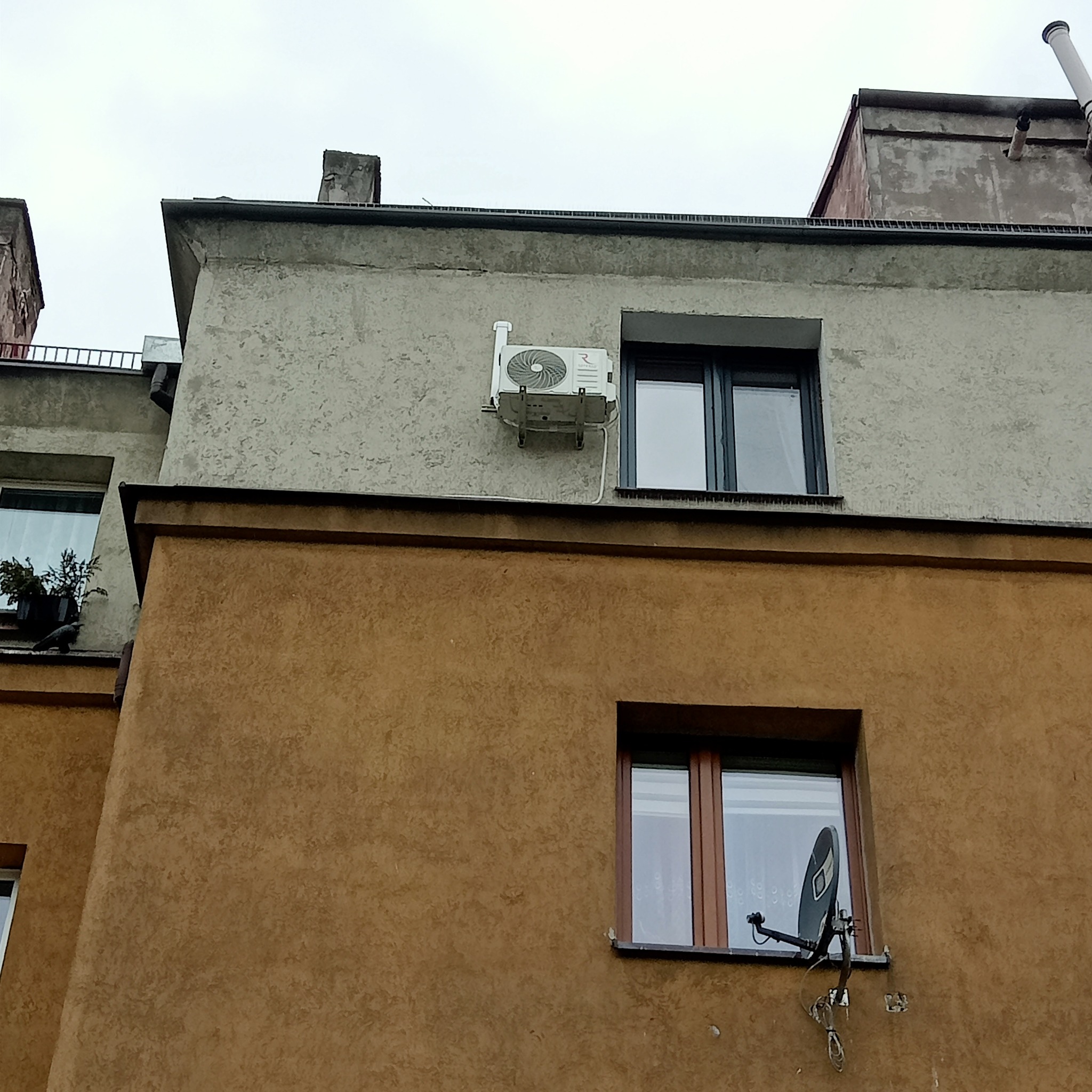 Zewnętrzna jednostka klimatyzacji zamontowana na elewacji budynku mieszkalnego, widoczna antena satelitarna na ścianie poniżej okna.