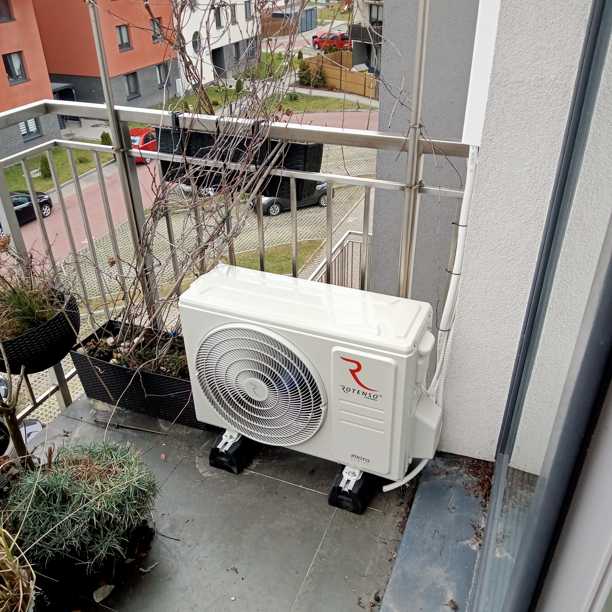 Zewnętrzna jednostka klimatyzacji Rotenso IMOTO zamontowana na balkonie w bloku mieszkalnym, widok z góry na jednostkę i fragment balkonu z donicami z roślinami.