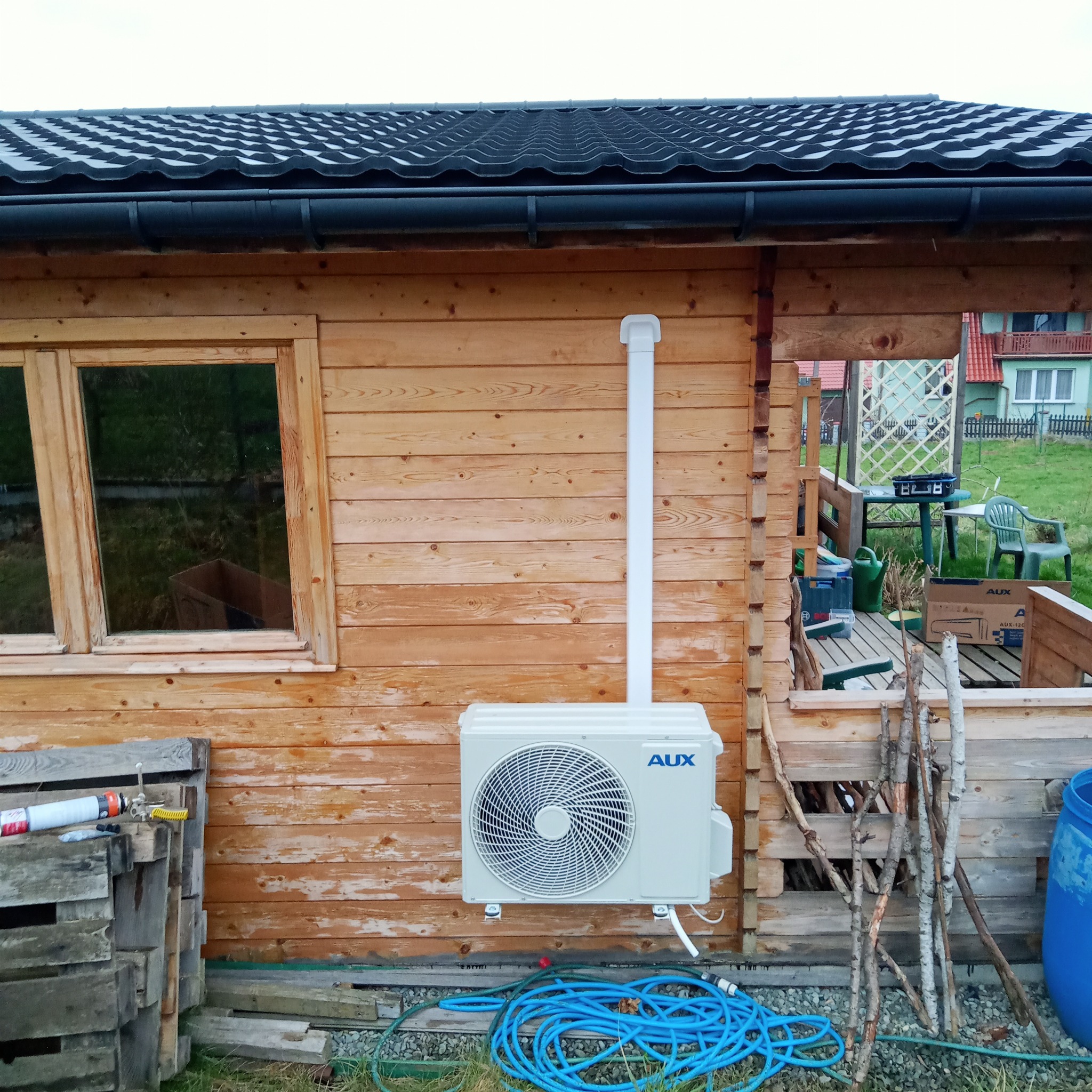 Zewnętrzna jednostka klimatyzacji AUX zamontowana na drewnianej ścianie domku letniskowego, z widocznymi elementami instalacji i niebieskim wężem ogrodowym na trawie.