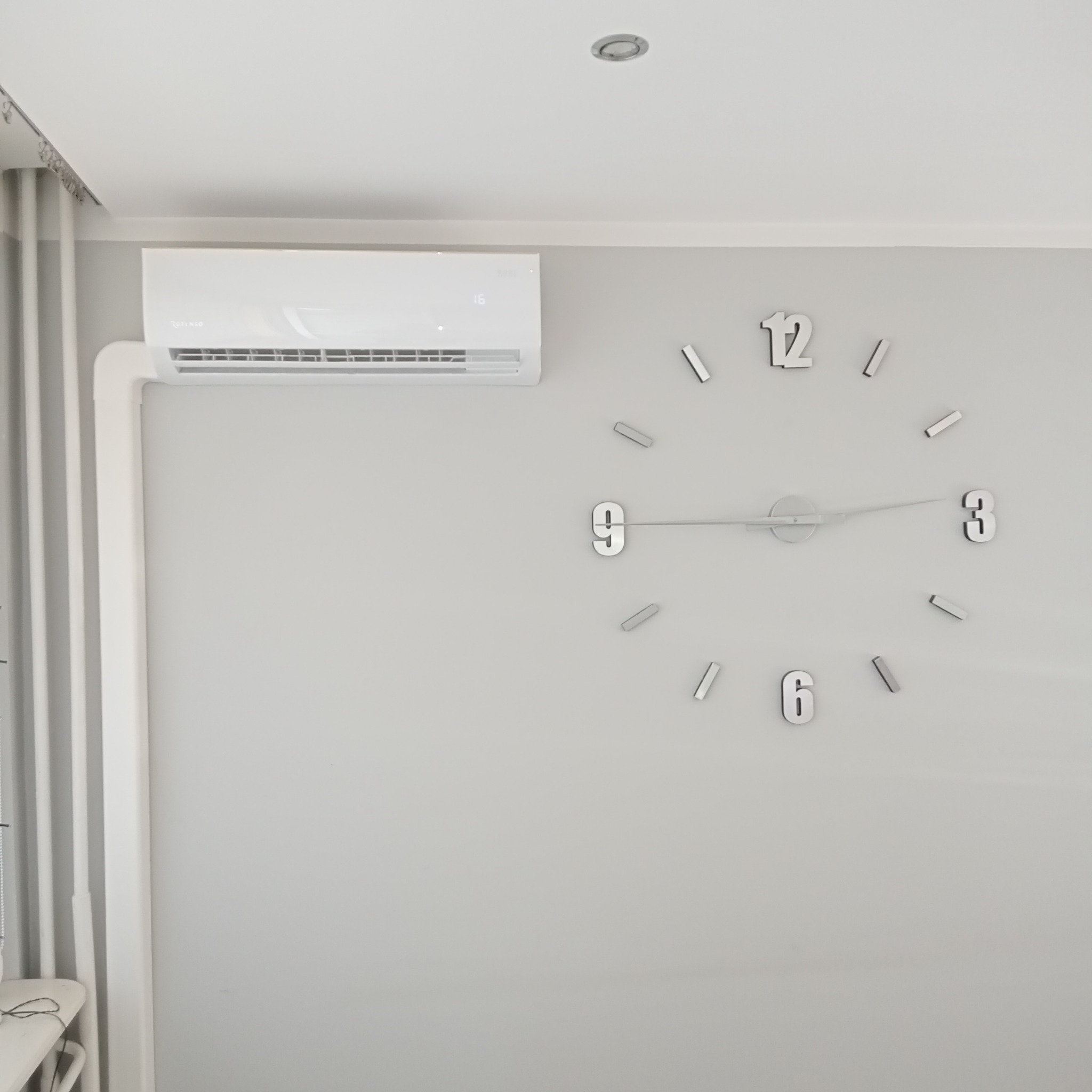 Biały klimatyzator na ścianie w pokoju z minimalistycznym, srebrnym zegarem ściennym.