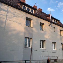 STAM - Ocieplony budynek wielorodzinny z szarą elewacją, brązowym dachem i białymi oknami, widoczne odbicia światła słonecznego na ścianie i cień drzewa.