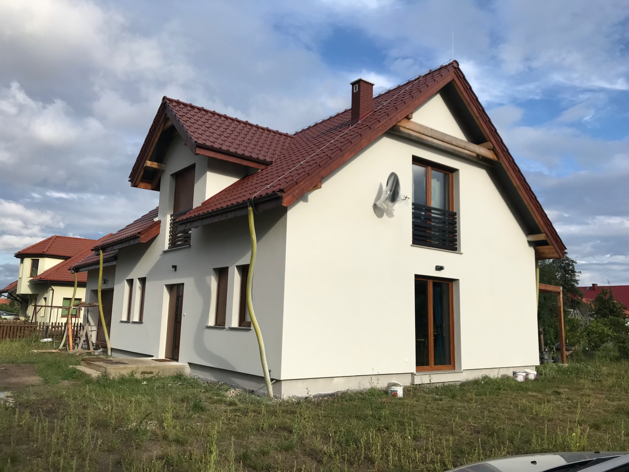 Świeżo otynkowany dom jednorodzinny z czerwoną dachówką, widoczna zielona rura do agregatu tynkarskiego i wiaderko z farbą przy ścianie