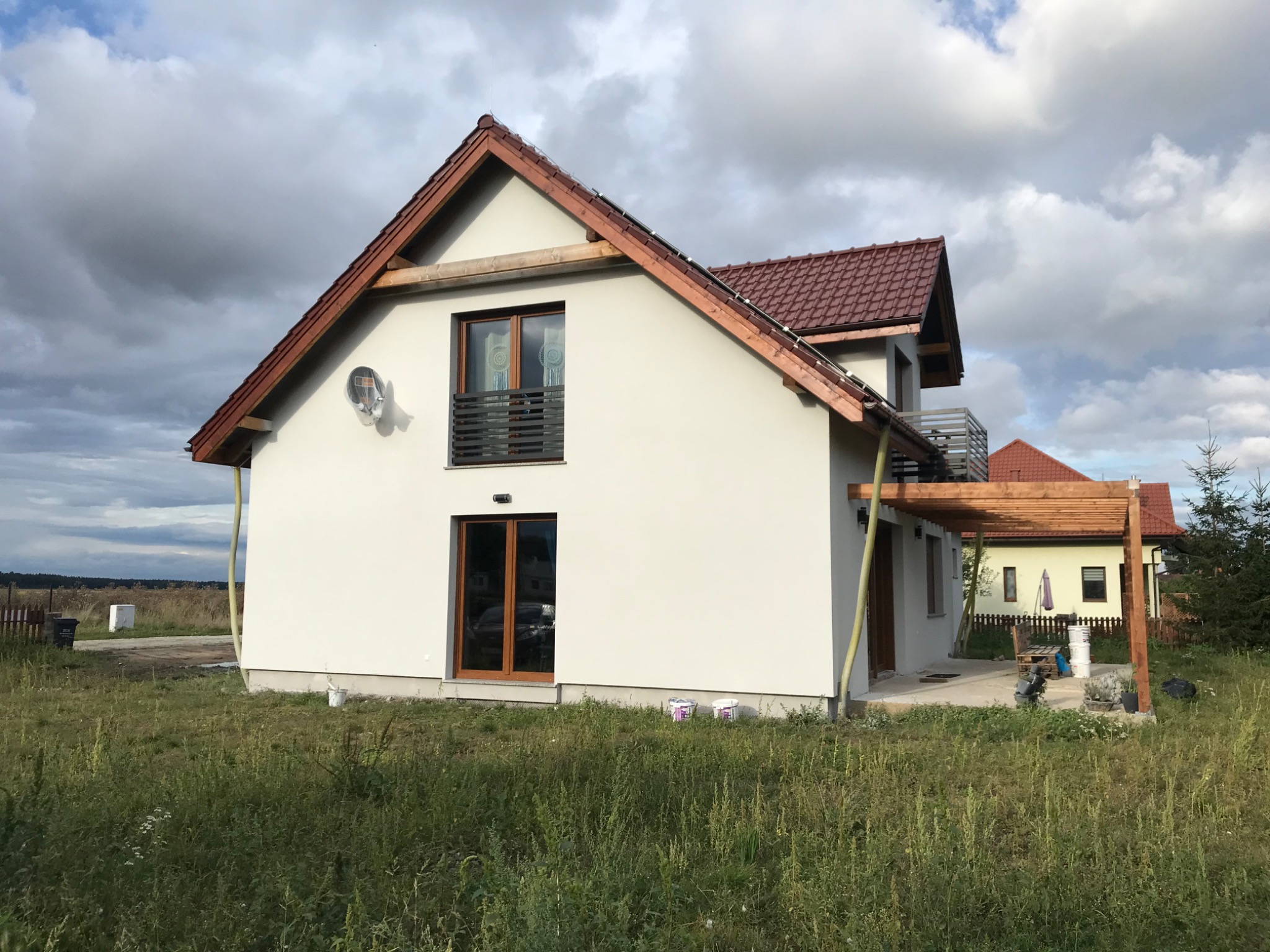 Nowo otynkowany dom jednorodzinny z brązową dachówką, drewnianą pergolą i anteną satelitarną na elewacji, otoczony wysoką trawą.