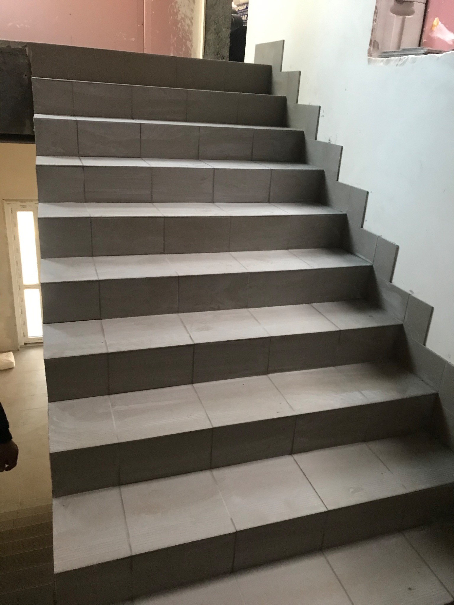 Schody wyłożone jasnymi, kwadratowymi płytkami ceramicznymi, widok z dołu, częściowo widoczna ściana po prawej stronie, w tle drzwi.