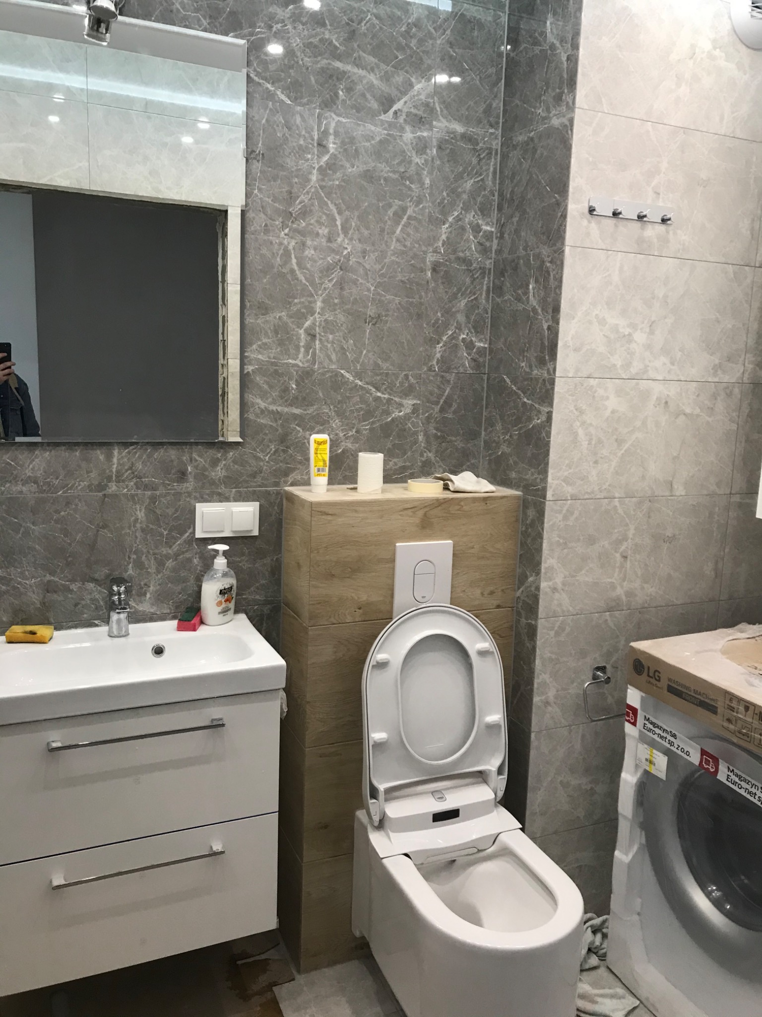 Nowoczesna łazienka z szarymi płytkami imitującymi marmur, białą umywalką z szafką, wiszącą toaletą z deską myjącą i nową pralką LG w kartonie, widoczne akcesoria łazienkowe.