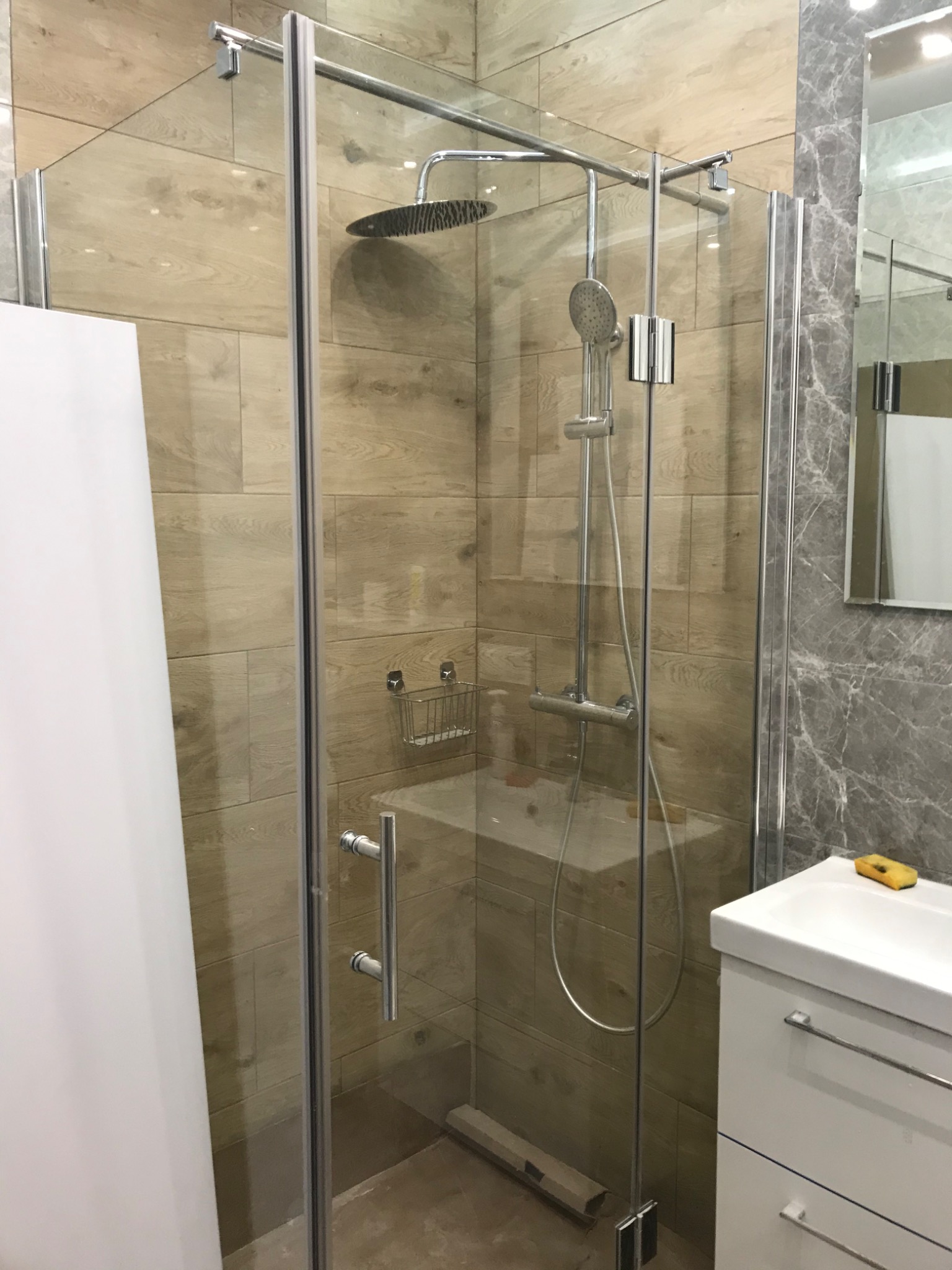 Kabina prysznicowa z deszczownicą i baterią termostatyczną na tle drewnopodobnych płytek w wykończonej łazience, widoczna umywalka z szafką i lustro.