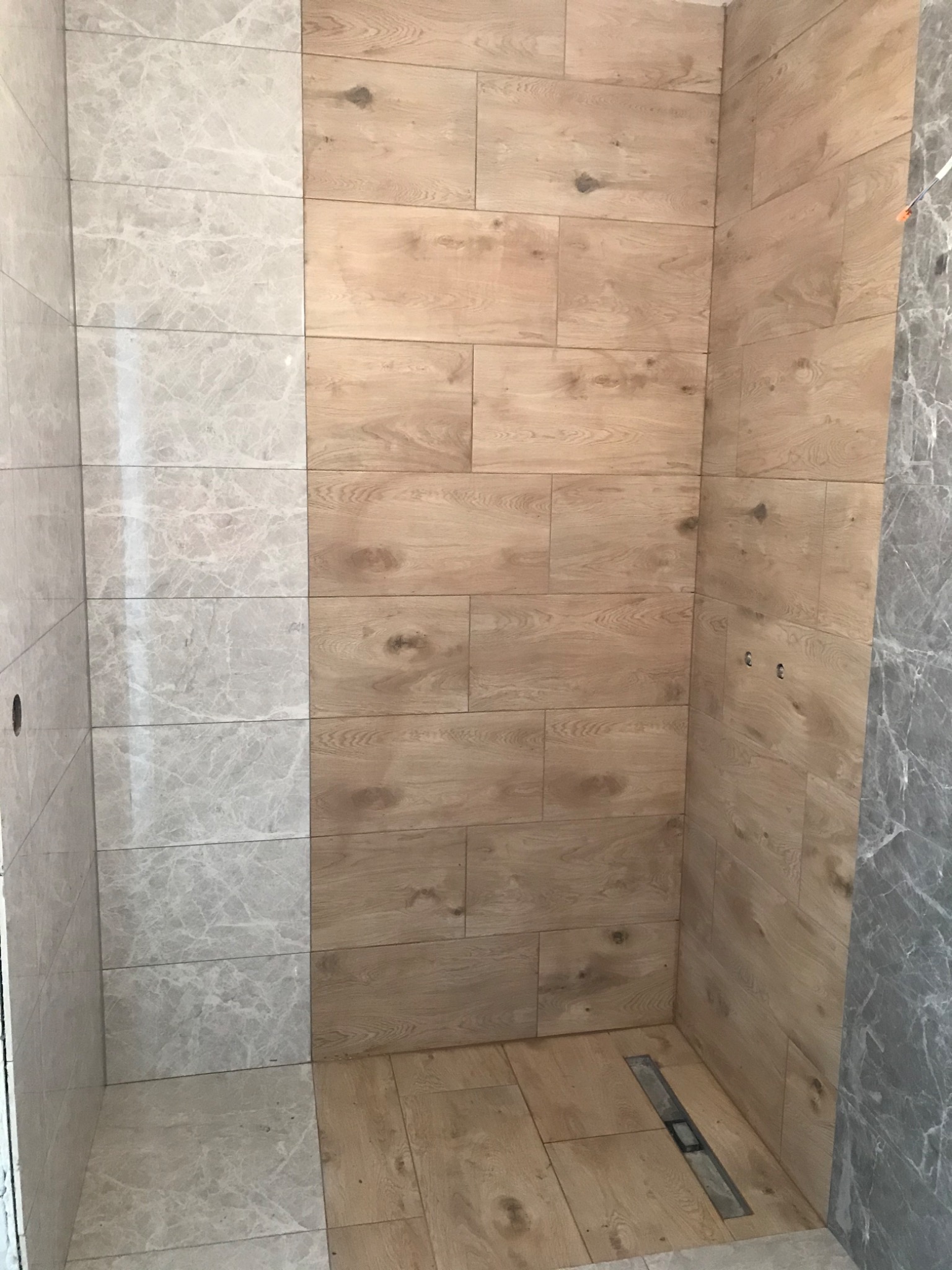 Wykończony prysznic z dwiema ścianami wyłożonymi płytkami imitującymi drewno, trzecią ścianą z szarych płytek imitujących marmur i odpływem liniowym w podłodze.