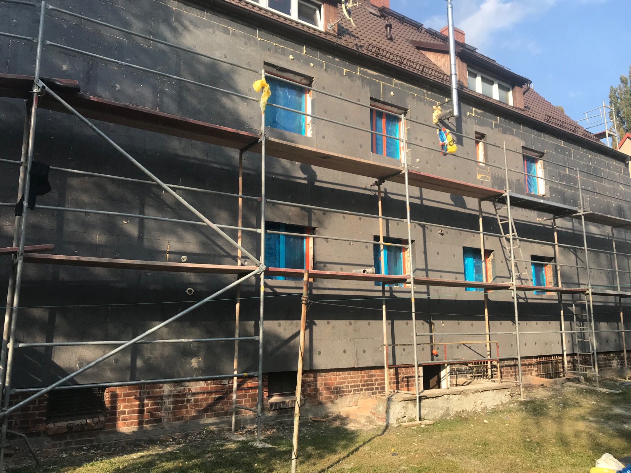 Budynek wielorodzinny w trakcie ocieplania, widoczne rusztowania, styropian na elewacji i okna zabezpieczone folią.