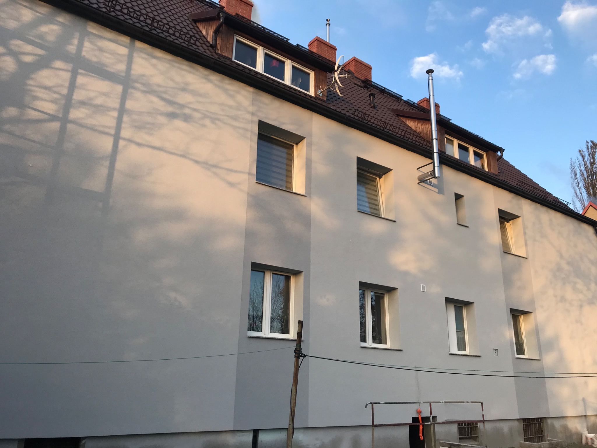 Ocieplony budynek wielorodzinny z szarą elewacją, brązowym dachem i białymi oknami, widoczne odbicia światła słonecznego na ścianie i cień drzewa.