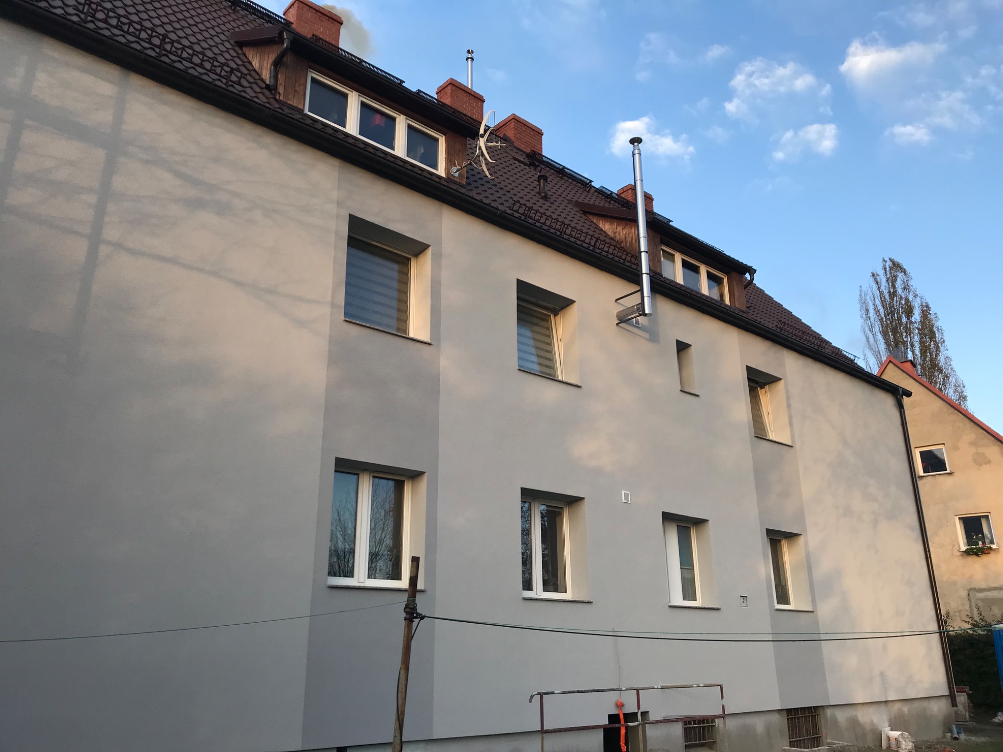 Ocieplony budynek wielorodzinny z szarą elewacją, widoczne kominy i instalacje na dachu, fragment rusztowania u dołu.