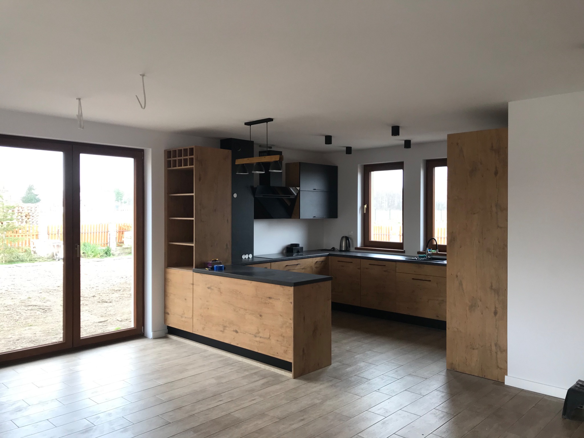 Nowoczesna, jasna kuchnia z drewnianymi frontami szafek i czarnymi akcentami, widok na ogród przez duże okno balkonowe, podłoga wyłożona płytkami imitującymi drewno, surowe haczyki montażowe...