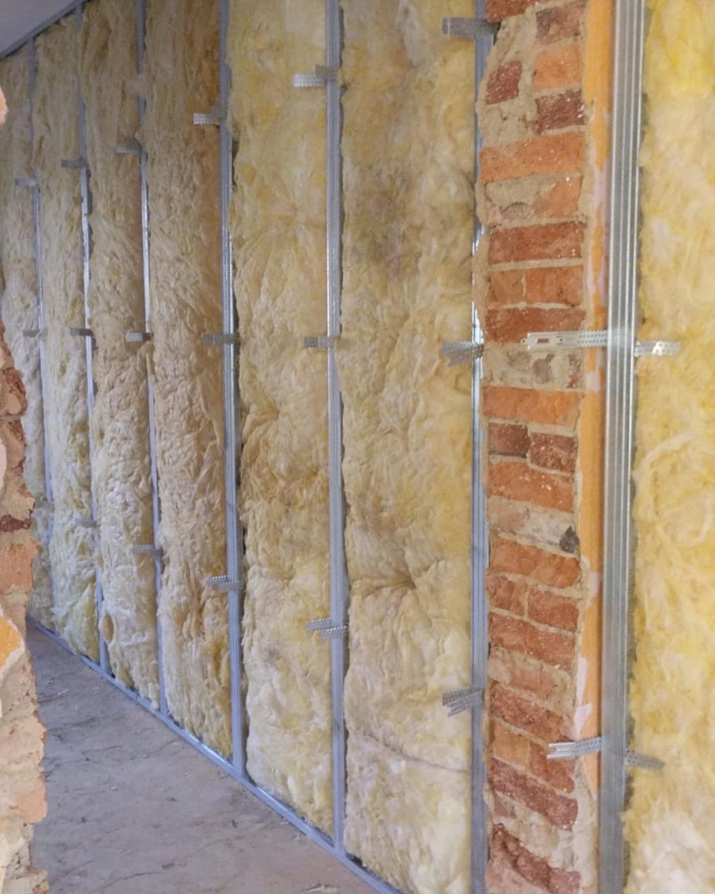 Ocieplenie ściany wełną mineralną, widoczne profile metalowe i fragment muru z cegły, etap prac wykończeniowych.