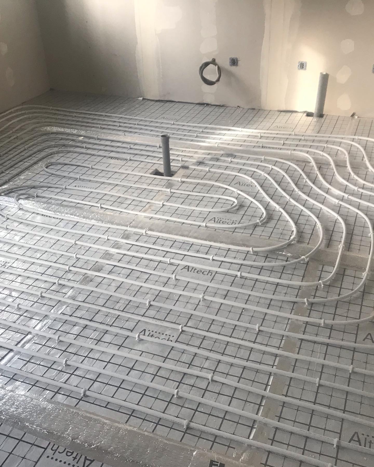 Rozłożone rury ogrzewania podłogowego Altech na srebrnej folii z czarną siatką w surowym pomieszczeniu przed wylewką, widoczne pionowe rury instalacyjne.