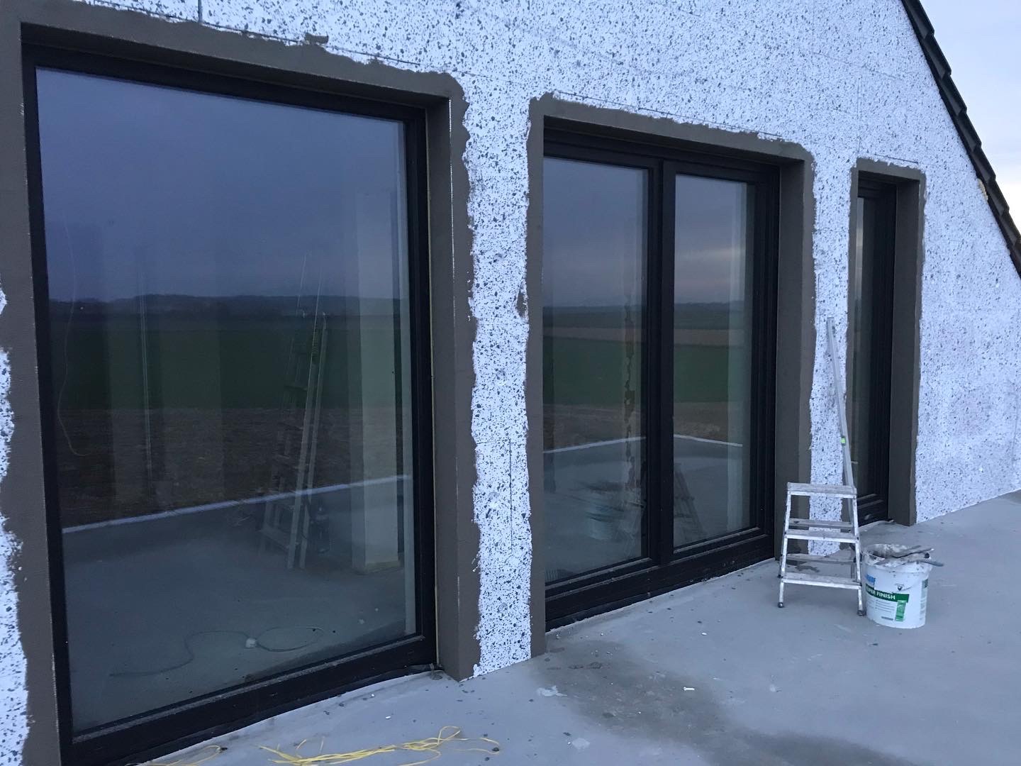 Dom w trakcie ocieplania, widoczne okna z obramowaniem w surowym tynku, drabina i wiadro z zaprawą na zewnątrz budynku.