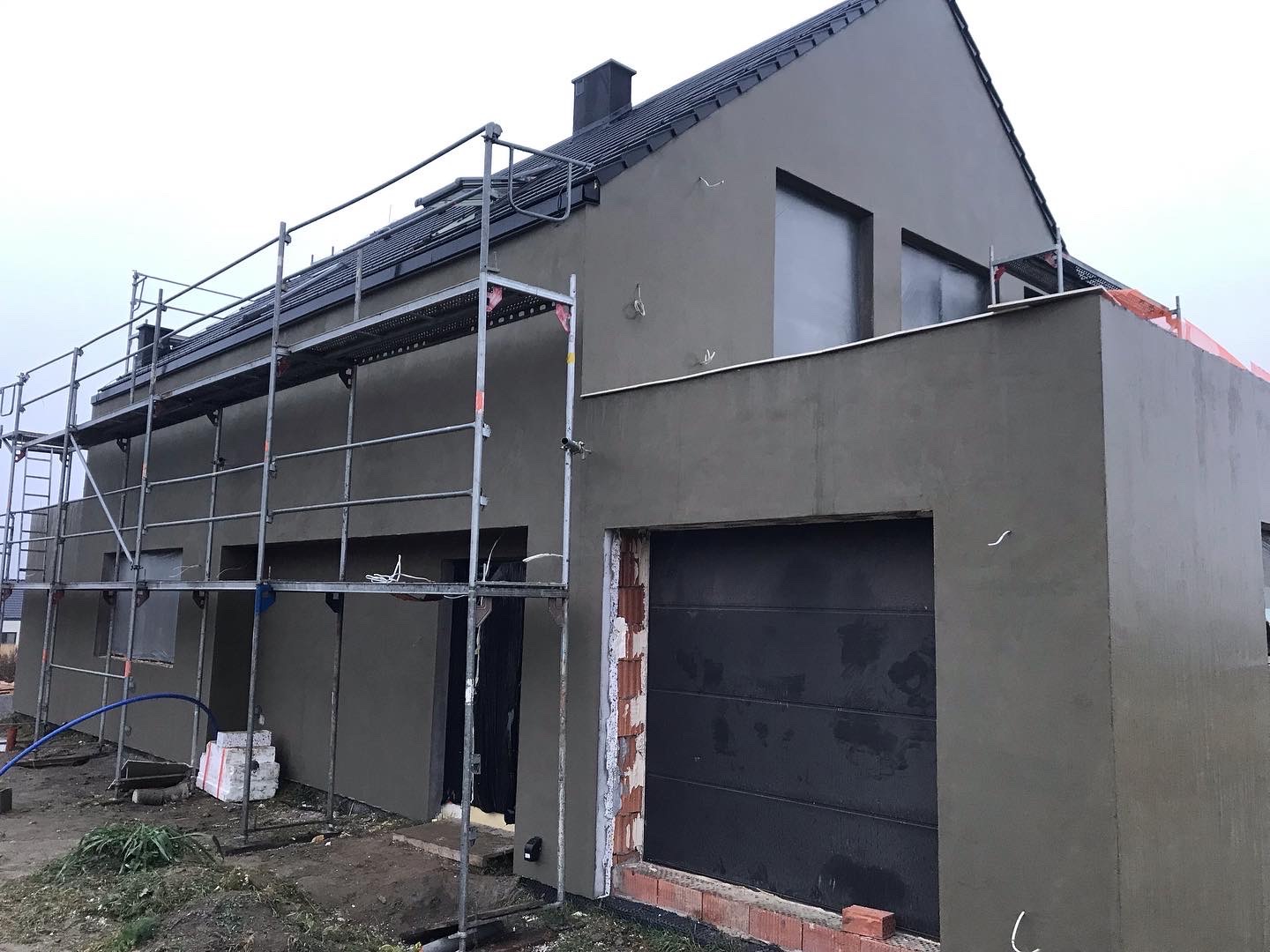 Dom jednorodzinny w trakcie ocieplania, widoczne rusztowania i świeżo nałożony tynk w odcieniach szarości, brama garażowa w kolorze antracytowym, przygotowane materiały budowlane na ziemi.