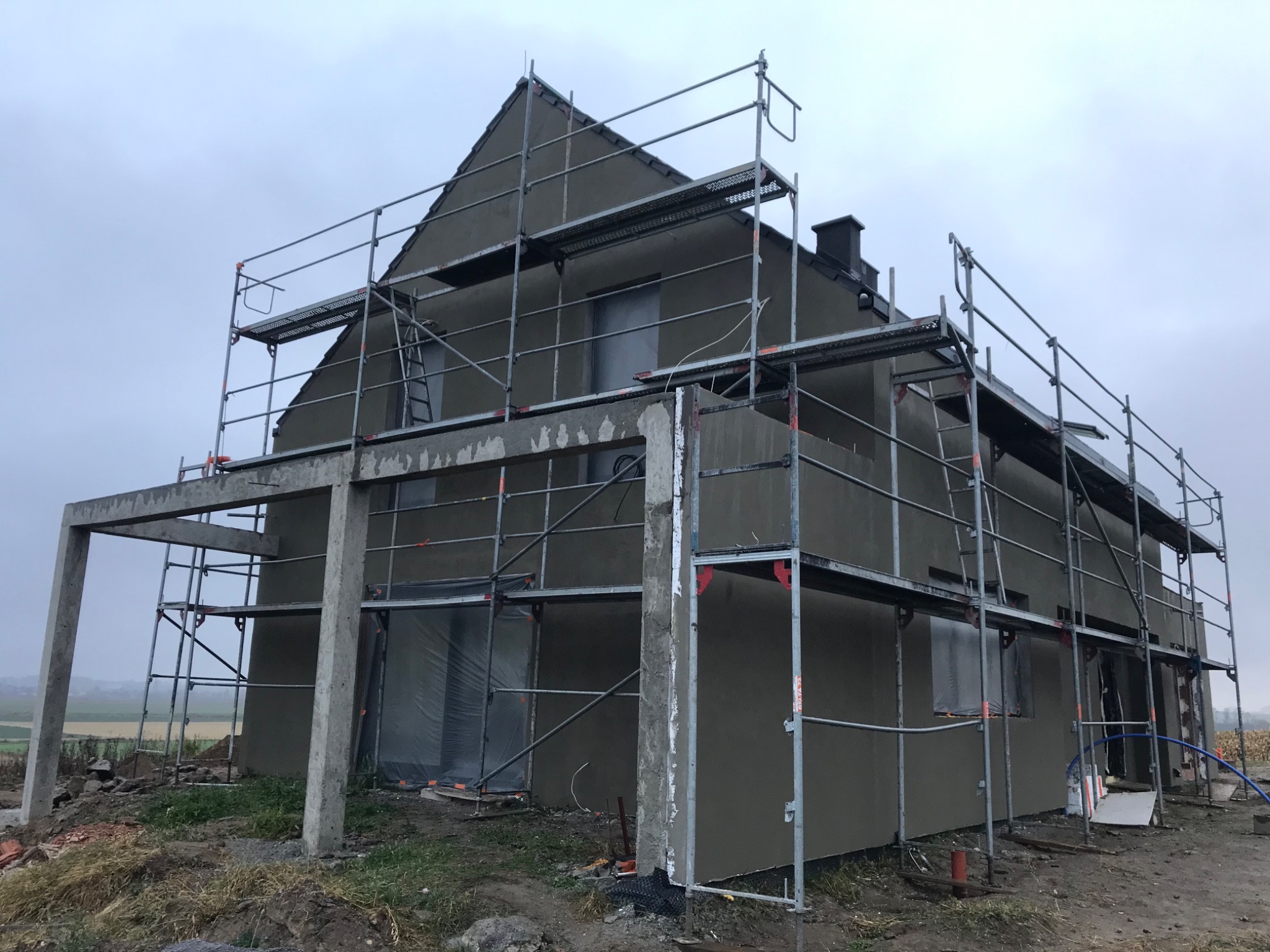 Dom jednorodzinny w trakcie ocieplania, z widocznymi rusztowaniami i świeżo nałożoną warstwą izolacyjną, okna zabezpieczone folią, pochmurne niebo w tle.