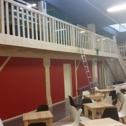 Green Bottles - Dwupoziomowe wnętrze biurowe w stylu stodoły z czerwonymi ścianami, drewnianymi balustradami i schodami, oświetlone lampami sufitowymi i naturalnym światłem, wyposażone w proste drewniane stoły...