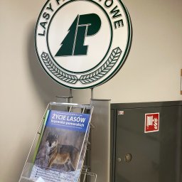Green Bottles - Logo Lasów Państwowych nad stojakiem z broszurami 'Życie Lasów kujawsko-pomorskich' i szafką hydrantową w tle.