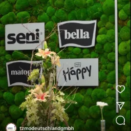 Żywa ściana z mchu, dekorowana kompozycją kwiatową i tabliczkami z logo marek Seni, Bella, Matopat i Happy.