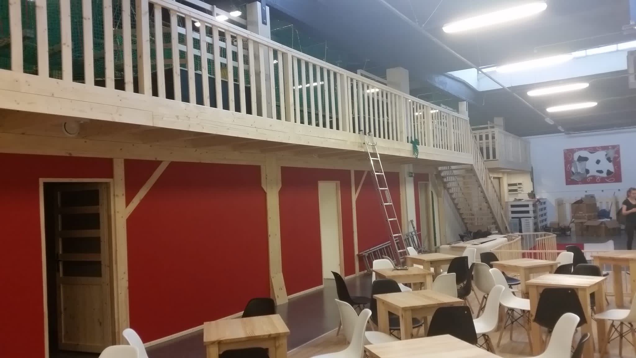 Dwupoziomowe wnętrze biurowe w stylu stodoły z czerwonymi ścianami, drewnianymi balustradami i schodami, oświetlone lampami sufitowymi i naturalnym światłem, wyposażone w proste drewniane stoły...