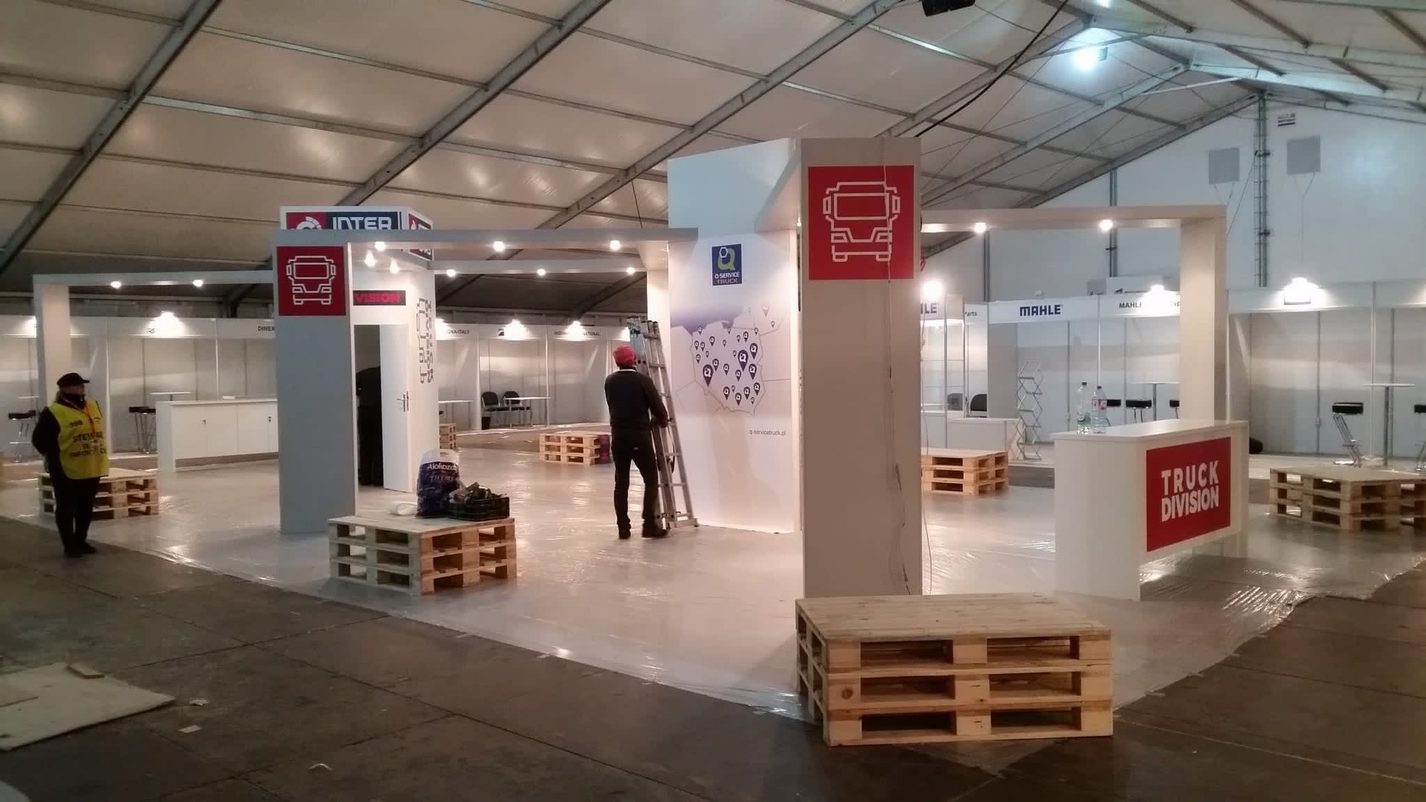 Montaż stoiska targowego: mężczyzna na drabinie instaluje grafikę z logo firmy Q Service Truck, w tle inne stoiska z logo Inter, Mahle, na podłodze folia ochronna i palety.