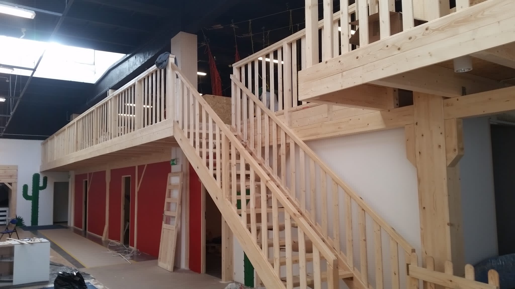 Wnętrze biura lub sali zabaw z drewnianą antresolą, schodami i balustradami, w stylu western, z czerwonymi ścianami i zielonym kaktusem.