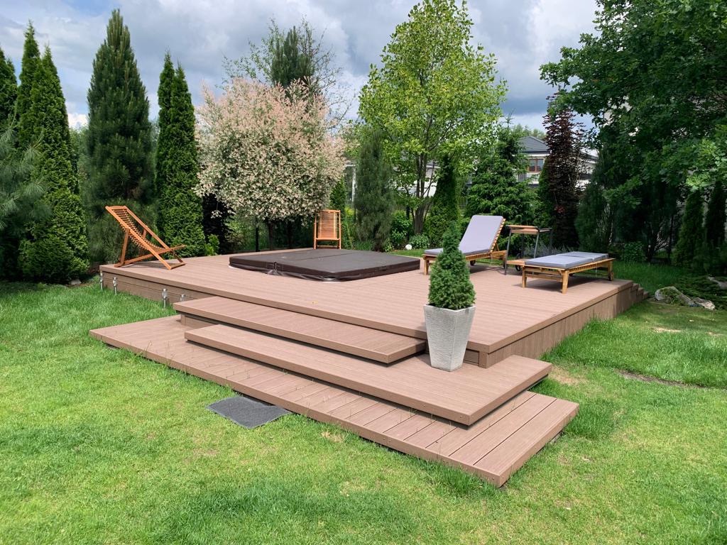 Drewniany taras z jacuzzi i leżakami w otoczeniu zieleni, Toruń. Kompozycja uzupełniona o rośliny w donicach i ozdobne drzewa.