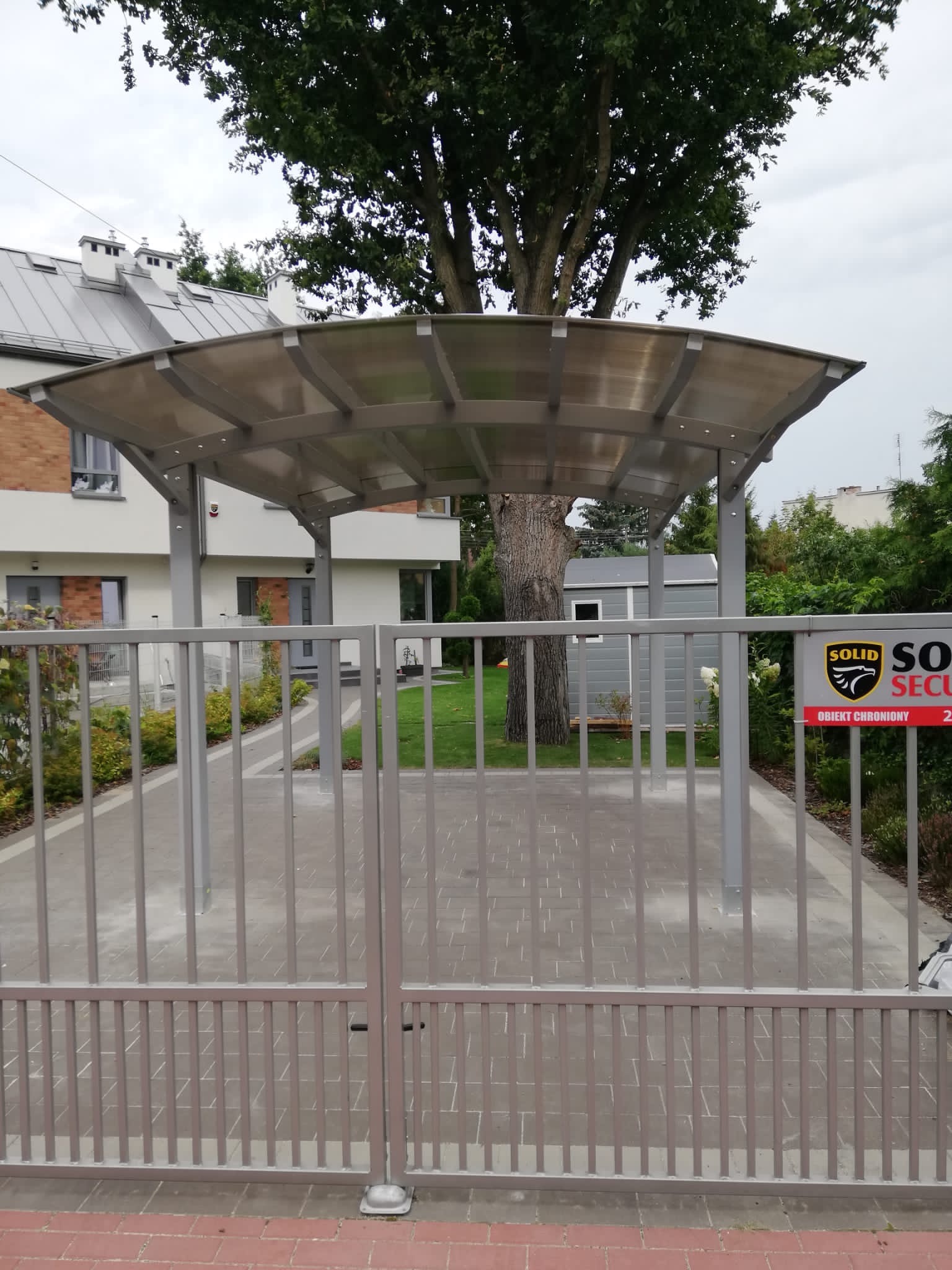 Szara metalowa brama wjazdowa z zadaszeniem z poliwęglanu, widoczny budynek mieszkalny w tle i tabliczka informacyjna o ochronie obiektu.