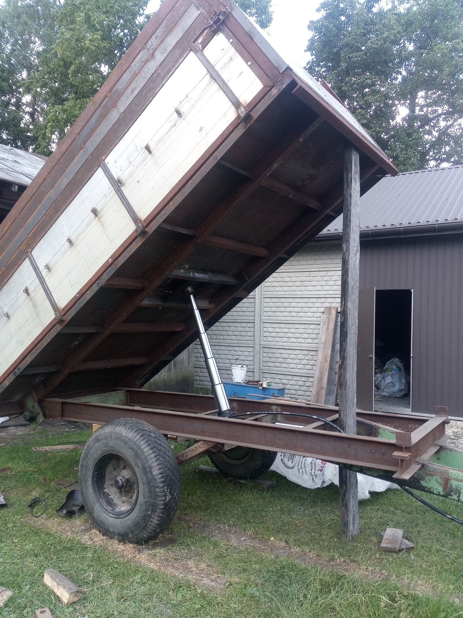 Przyczepa rolnicza z podniesioną skrzynią ładunkową, widoczny siłownik hydrauliczny, opony i metalowa konstrukcja, na trawiastym podłożu, w tle budynek gospodarczy.