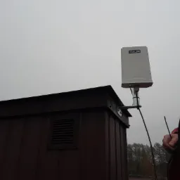 Biała antena z logo na dachu brązowego budynku z kratkami wentylacyjnymi, na tle szarego nieba i drzew, z fragmentem osoby w czerwonej kurtce po prawej stronie.