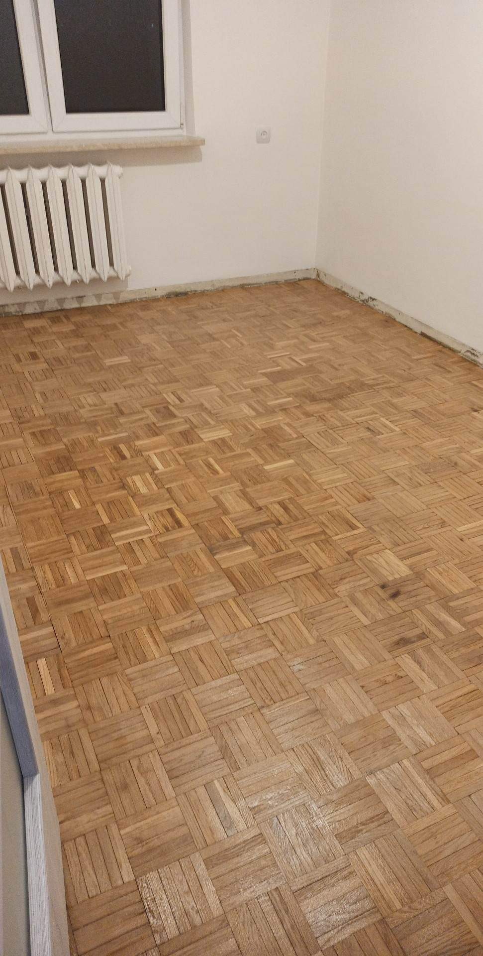 Ułożona parkietowa podłoga w pomieszczeniu z białymi ścianami, widoczny grzejnik pod oknem.