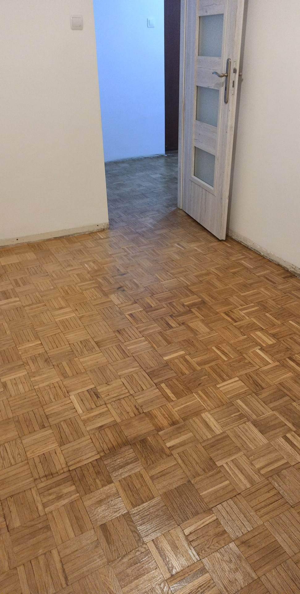 Parkiet ułożony w kwadraty w pustym pokoju z otwartymi drzwiami i pomalowanymi na biało ścianami.