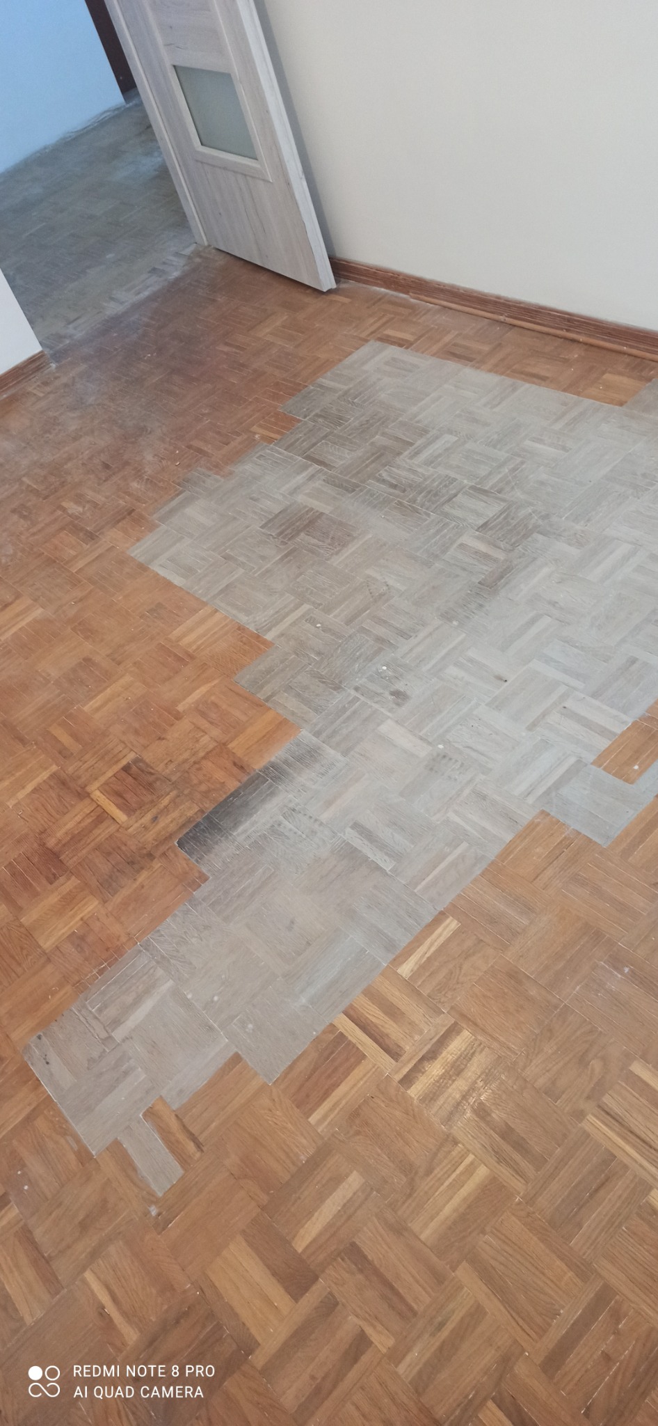 Fragment podłogi parkietowej w jodełkę, częściowo wyszlifowany, z widoczną różnicą w kolorze i fakturze, drzwi w tle.